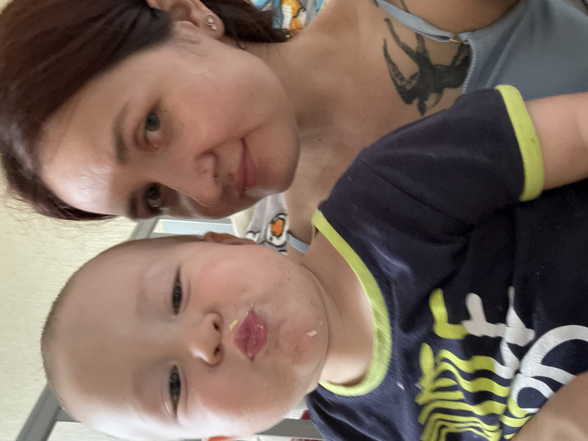 The image shows a close-up selfie of a young child and a woman. The child, facing the camera, has their lips pursed and food residue around their mouth. They are wearing a dark blue t-shirt with a neon green trim and a graphic design. To the right, the woman is also looking at the camera, with her hair pulled back. She is wearing a light blue top and has a tattoo of a bird on her upper chest. The background is blurred, suggesting an indoor setting, possibly a home. The specific location in Naberezhnye Chelny, Russia, cannot be determined from the visual information provided.