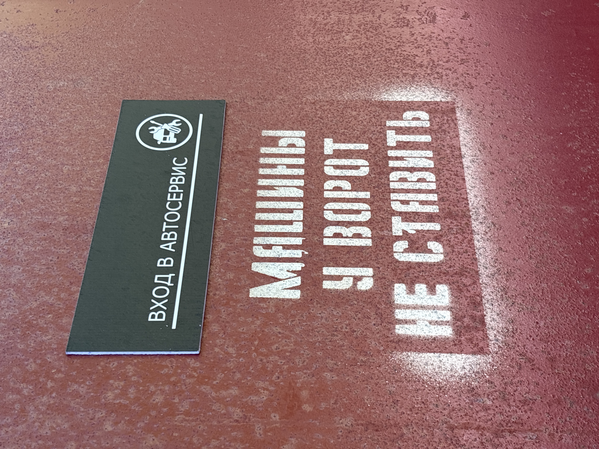 The image shows a sign on a red surface in Chaikovsky, Russia. A dark gray rectangular sign on the left reads "ВХОД В АВТОСЕРВИС" ("Entrance to Car Service") vertically, with a logo of a wrench and piston in a circle above the text. To the right, painted in white stencil letters on the red surface, is the phrase "МАШИНЫ У ВОРОТ НЕ СТАВИТЬ!" ("Do not park cars at the gate!").