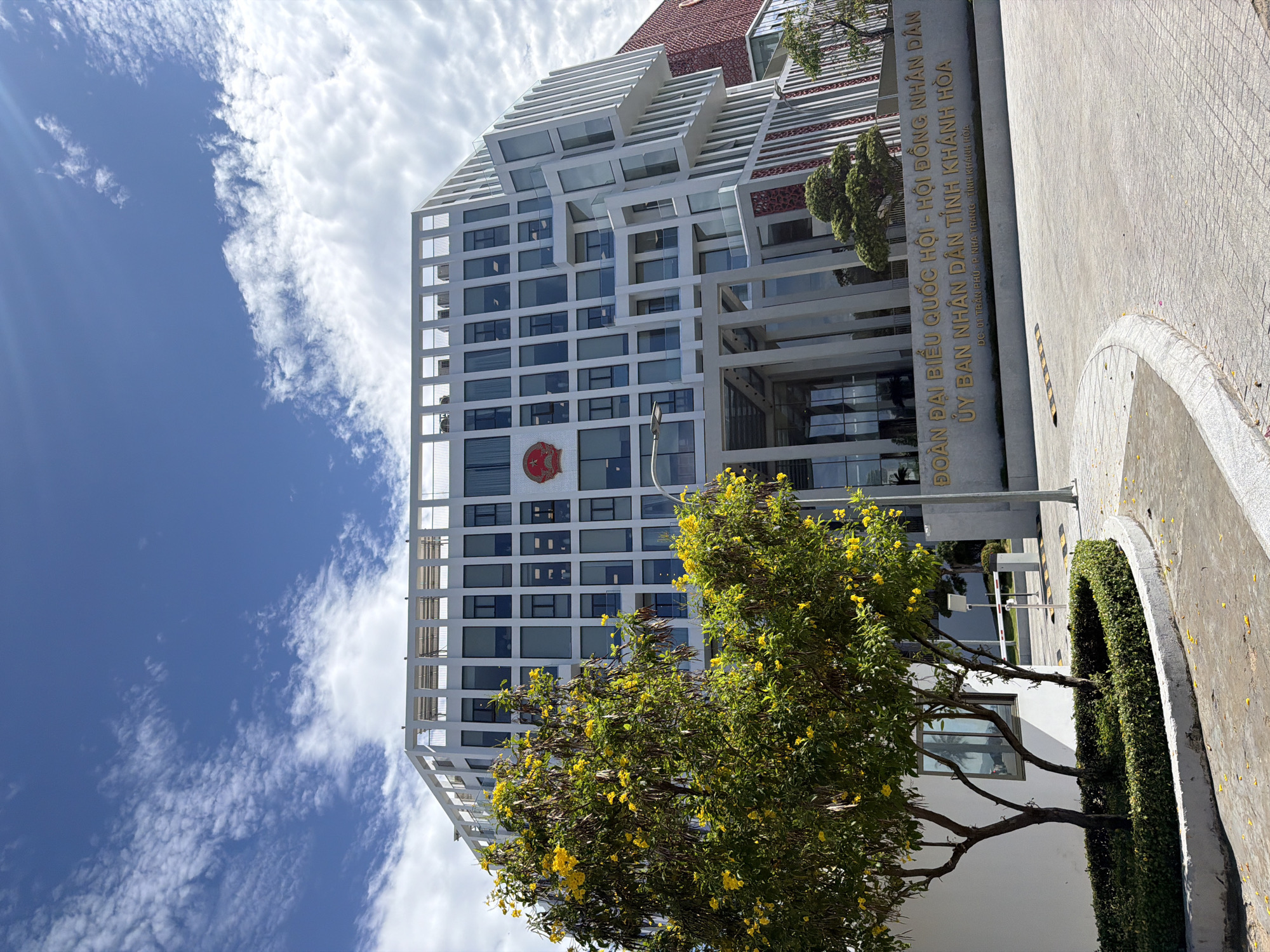 A modern, multi-story white building with a distinctive contemporary design stands prominently under a bright blue sky filled with white, wispy clouds. The building's facade features numerous rectangular windows, some dark-tinted, and a pattern of white architectural slats and frames. A red and gold emblem, likely representing a national or provincial authority, is visible on one of the building's lower sections. The upper levels of the building showcase a complex, tiered, and angular roofline.
In the foreground, a vibrant green tree with numerous yellow flowers partially obscures the lower part of the building. To the right of the building, a large stone sign displays text in Vietnamese, identifying the structure: "ĐOÀN ĐẠI BIỂU QUỐC HỘI - HỘI ĐỒNG NHÂN DÂN / ỦY BAN NHÂN DÂN TỈNH KHÁNH HÒA / ĐC: 01 TRẦN PHÚ P. NHA TRANG TỈNH KHÁNH HÒA". This translates to "Delegation of the National Assembly - People's Council / People's Committee of Khanh Hoa Province / Address: 01 Tran Phu, Nha Trang, Khanh Hoa Province."
The building is situated in Nha Trang Ward, Vietnam, in what appears to be a well-maintained public or governmental complex. The ground in front is paved, with a visible curb and manicured green hedges. The bright sun casts clear light, with sun rays visible in the upper left of the frame, indicating a sunny day. No people or specific activities are visible, presenting a serene, architectural landscape.