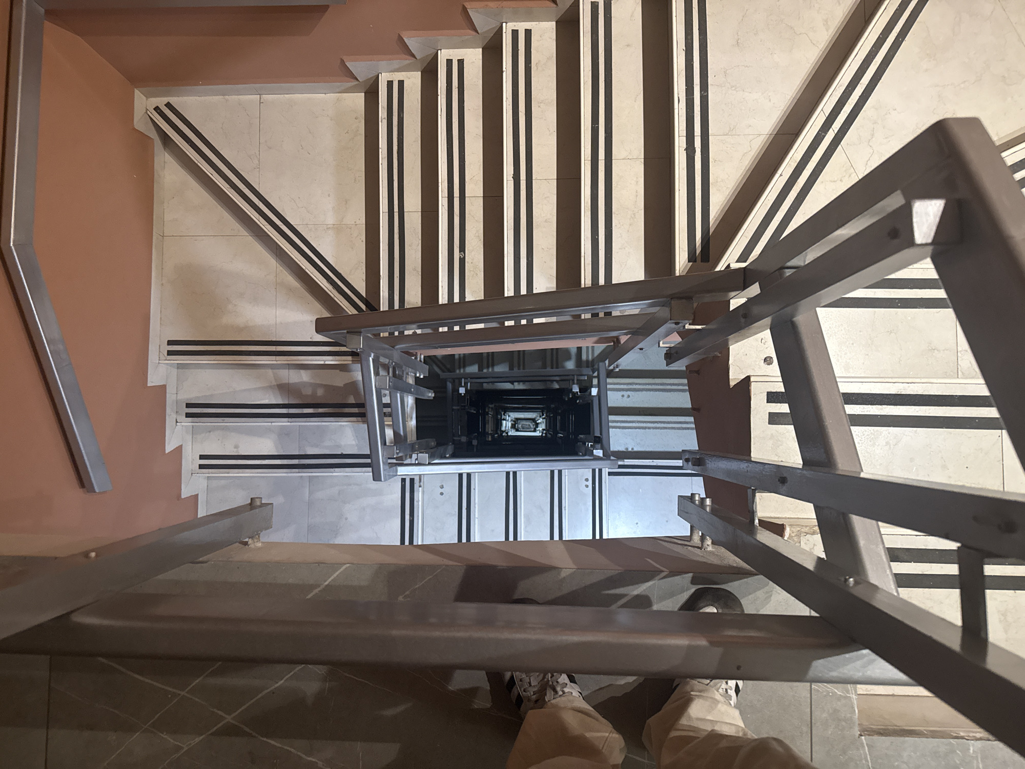 A high-angle, slightly distorted perspective depicts a stairwell in what appears to be Mexico City, Mexico. The staircase descends into a deep shaft, where the steps are lined with light-colored tiles. Each step has anti-slip stripes near the edge. A metal guardrail surrounds the stairwell. In the immediate foreground, a person stands with their feet visible, wearing shoes and light-colored pants. The person is positioned at the top of the stairs, looking down into the stairwell. The surrounding walls are pale orange.