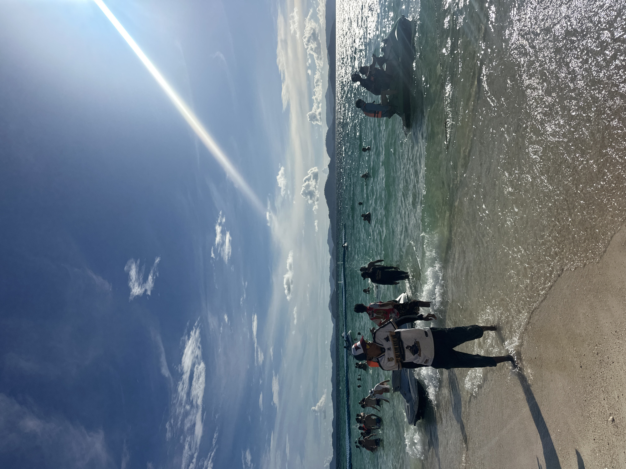 The image depicts a group of people on a sandy beach with clear, shallow ocean water. The sun is bright, casting strong highlights on the water and creating a visible sunbeam in the sky. The sky is a deep blue with scattered white clouds. In the foreground, a person is standing in the shallow water, wearing a white t-shirt with text, dark pants, and a helmet. Behind them, several other people are visible, some standing in the water and others near a dark boat. Further out in the water, more people and what appear to be jet skis or small boats can be seen. The overall scene suggests a sunny day at a tropical beach, likely a tourist destination, with recreational activities taking place. The time of day appears to be midday, given the brightness of the sun and the sharp shadows. The location cues, such as the clear water, sandy beach, and the presence of boats, are consistent with a beach setting in Phru Nai, Thailand. The emotions conveyed are generally positive, suggesting leisure and enjoyment of the outdoor environment.