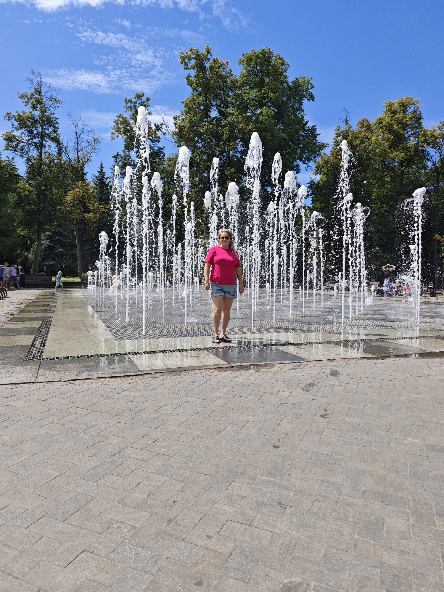 A woman stands in the middle of a fountain on a sunny day, with numerous water jets spraying upwards. She is wearing a pink t-shirt and denim shorts, and sunglasses. The fountain is located in a paved area with trees in the background. The sky is blue with scattered white clouds. The pavement is made of grey stone tiles laid in a herringbone pattern. In the distance, other people can be seen near benches and trees. The overall environment appears to be a park or public square in a city.