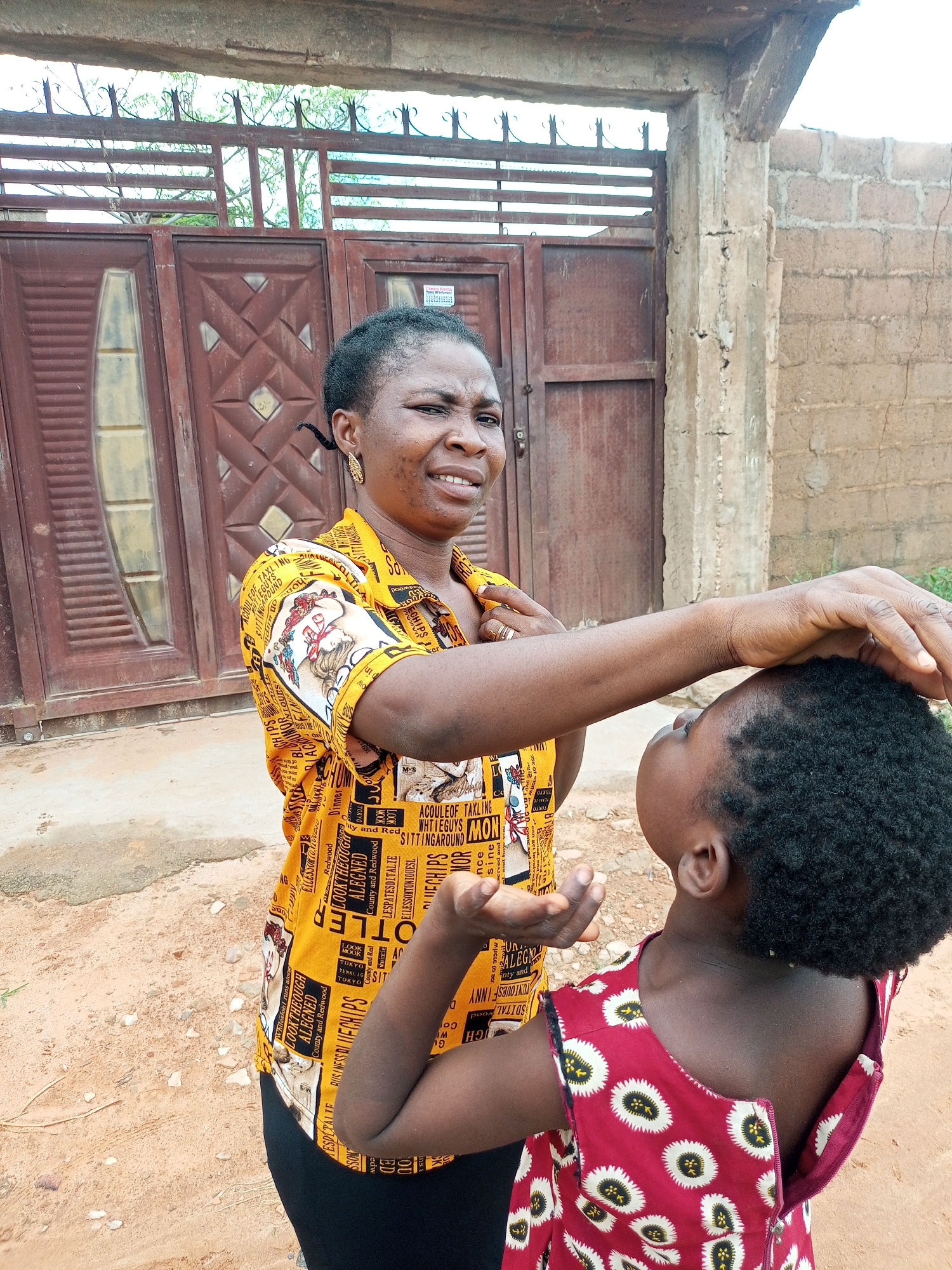 A medium shot captures two individuals outdoors in what appears to be an urban environment in Tempure, Nigeria. The foreground is dominated by the upper torso of a child facing away from the camera, wearing a red dress with a white and black floral pattern and a zipper down the back. The child's dark, curly hair is visible. To the left and slightly behind the child stands an adult woman, facing the child. She wears a yellow button-up shirt with a repeating black text and image pattern. Her right hand is gently placed on the child's head, and her facial expression is one of concern or perhaps mild admonishment. The background features a weathered, reddish-brown double door with ornate geometric patterns, set within a concrete structure. A chain-link fence with vertical bars is visible above the door. To the right of the door, a rough, textured brick wall forms the boundary of the scene. The ground is a sandy, reddish-brown dirt. The lighting suggests it is daytime, with soft, diffused light.