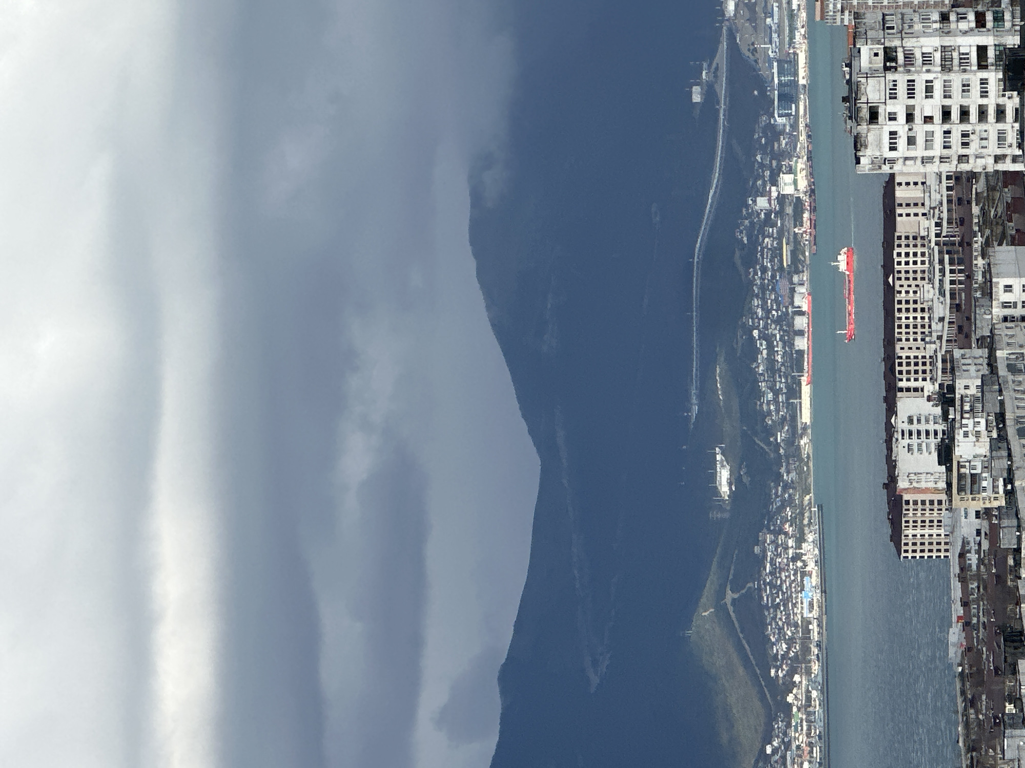 This image depicts a city in an unknown location, adjacent to a body of water, possibly a sea or harbor. A mountain looms in the background, partially obscured by clouds. The city is characterized by densely packed buildings. A red cargo ship is visible in the waterway, positioned between the city and the coastline opposite. A road or path runs along the lower portion of the mountain. The sky is predominantly overcast.