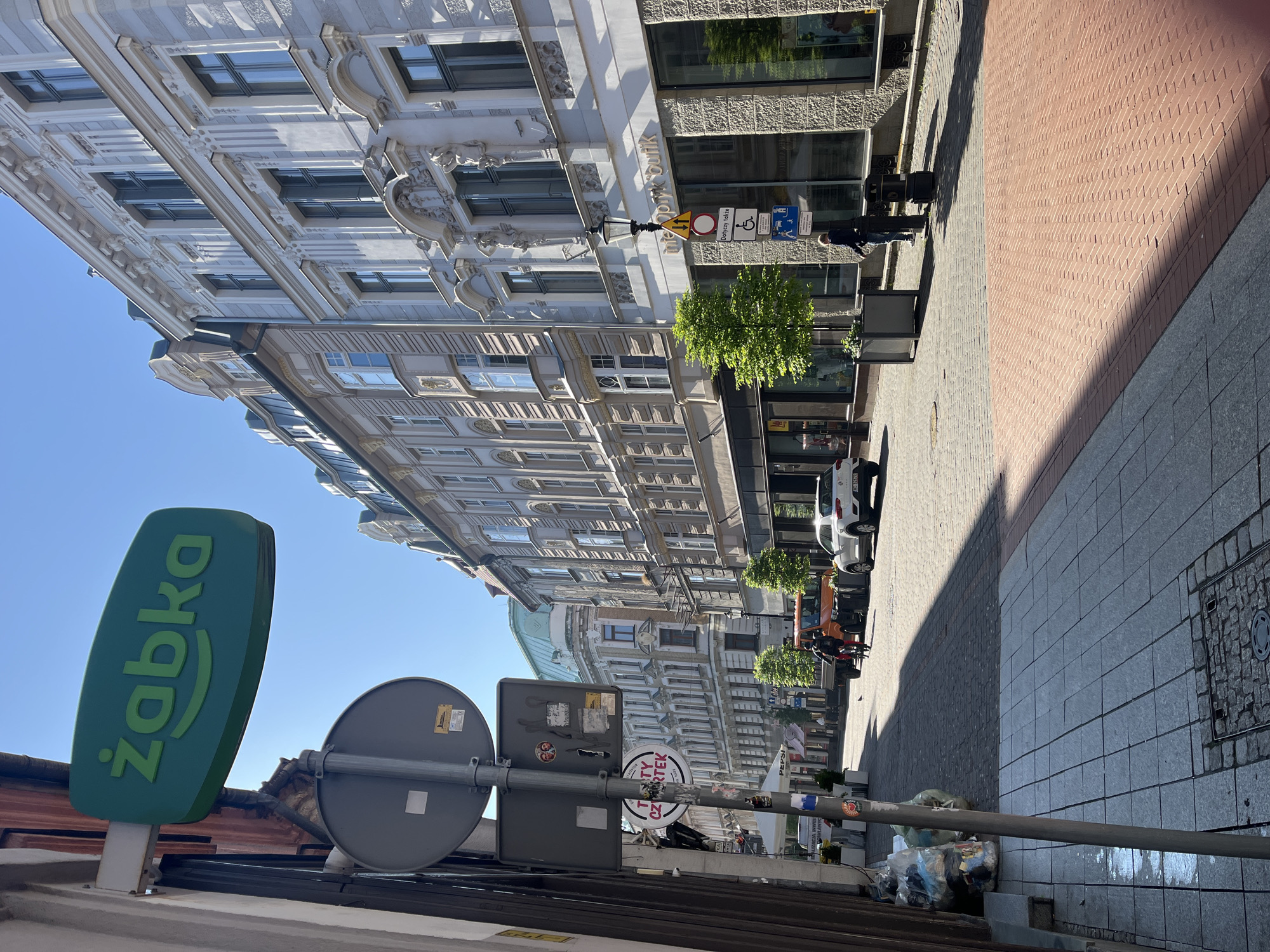 The image shows a street scene in an unidentified urban area. The view is from the edge of a sidewalk, looking down the street.
The street is lined with buildings on both sides. The architecture is mixed, featuring both older, ornate facades with detailed moldings and more modern glass and steel structures. There are signs indicating a "Żabka" convenience store, and other various commercial signs.
There are people visible on the sidewalk, including one individual walking. Cars and other vehicles are parked along the street. Street trees are planted along the sidewalk, and there are street lights and signs posted.
The sidewalk is paved with a combination of brick and gray stone blocks. There is a shadow cast across the sidewalk and street. There is some trash visible near the curb.
