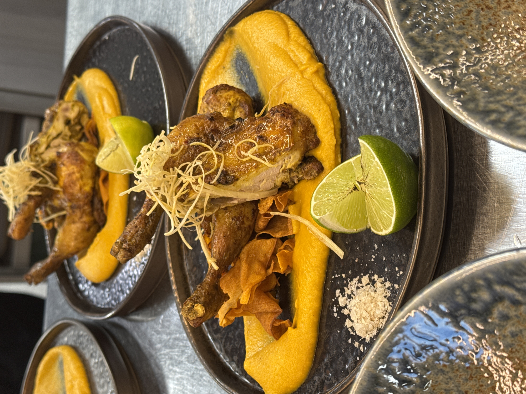 This image displays three plates of roasted chicken served on a bright orange purée, garnished with crispy fried shreds and lime wedges. Two plates are in focus, and a third is partially visible in the background. The plating is elegant, with a rustic charm. The chicken is golden brown and appears juicy. The orange purée is smooth and vibrant, providing a stark contrast to the dark plates. Crispy sweet potato chips are scattered around the chicken. A sprinkle of coarse sea salt is visible on the plate. The setting appears to be a restaurant kitchen or a food preparation area, with stainless steel surfaces visible. The image does not provide any information that would allow for the identification of a specific location in Sofia, Bulgaria.