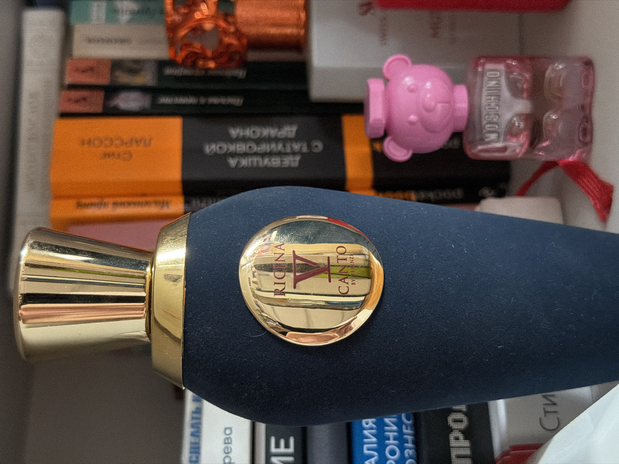 The image shows a close-up of a perfume bottle resting amongst several books and other objects. The bottle is dark blue with a gold-colored nozzle and a gold circular emblem bearing the brand name "RICINA CANTO BY TERENZI". Adjacent to the perfume bottle are several books, with titles in Cyrillic, including "Стиг Ларссон" (Stieg Larsson) and "ДЕВУШКА С ТАТУИРОВКой ДРАКОНА" (The Girl with the Dragon Tattoo). A pink toy bear (presumably a Moschino toy) is visible behind the perfume bottle. The arrangement suggests the objects are placed within a drawer or on a shelf. There is no indication in the image of its location being Moscow, Russia.