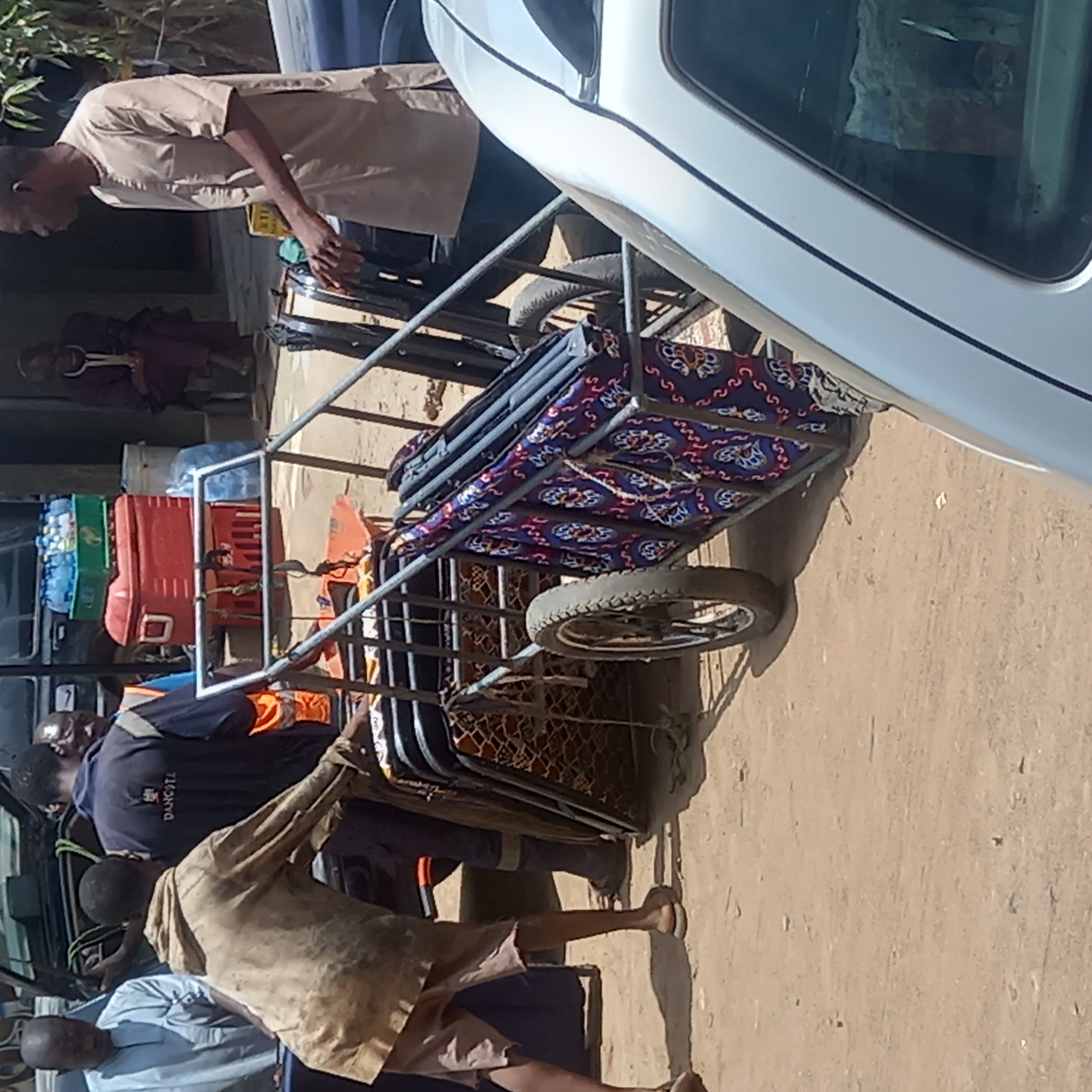 The image shows a makeshift hand-pulled cart parked beside a white van or truck on a dirt-covered street in Dadi Riba, Nigeria. The cart, constructed from metal tubing, has a cushion with a colorful patterned cover on top. It is loaded with items including water bottles, a red toolbox, and other miscellaneous objects. Several people are present. One man, wearing a tan shirt, stands near the front of the cart. Other individuals are nearby, one wearing an orange safety vest over a dark shirt. The logo "DANCOLT" is visible on the vest. The environment appears to be a roadside in a residential area. The image is taken during daylight, and the orientation of the photo is rotated ninety degrees clockwise.