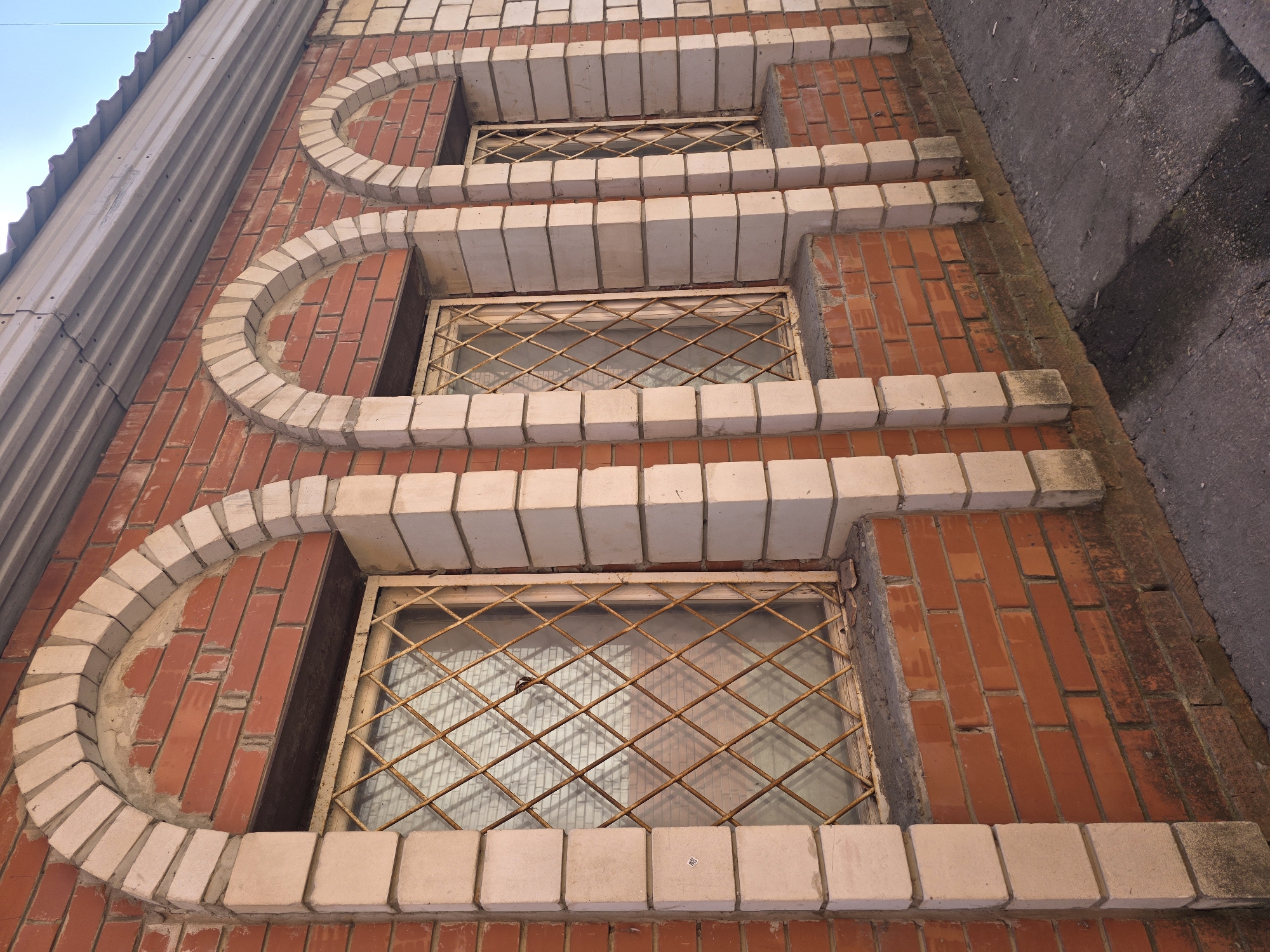 The photograph shows a section of a brick building facade, likely in Chaikovsky, Russia. The wall is primarily constructed of red bricks and features three windows. Each window has a decorative, semi-circular arch made of white bricks above it. The windows are protected by a diamond-patterned metal grid. The wall runs diagonally upwards, and the upper right corner of the image shows a section of concrete wall. To the left of the brick wall is a corrugated metal panel. The sky is partially visible in the upper left corner.
