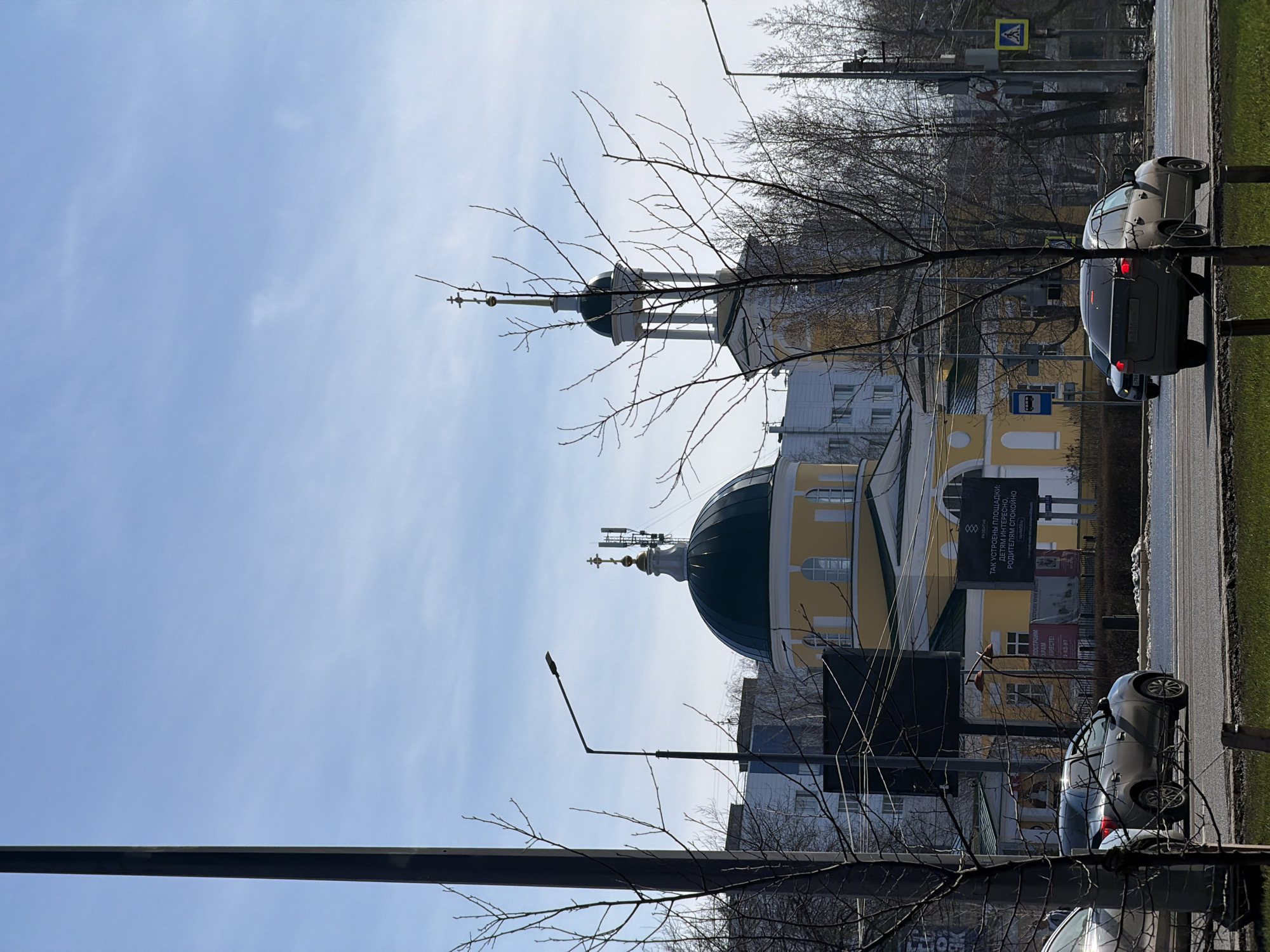 The image captures a street scene in Perm, Russia, focusing on a yellow and white church building with a prominent green dome. The church is partially obscured by bare tree branches in the foreground. A street with two cars parked along the curb occupies the right side of the frame. A bus stop sign and a pedestrian crossing sign are visible near the parked cars. A billboard with text in Russian stands adjacent to the church. The sky is blue with faint clouds, suggesting a sunny day. A lamppost stands near the building on the left.