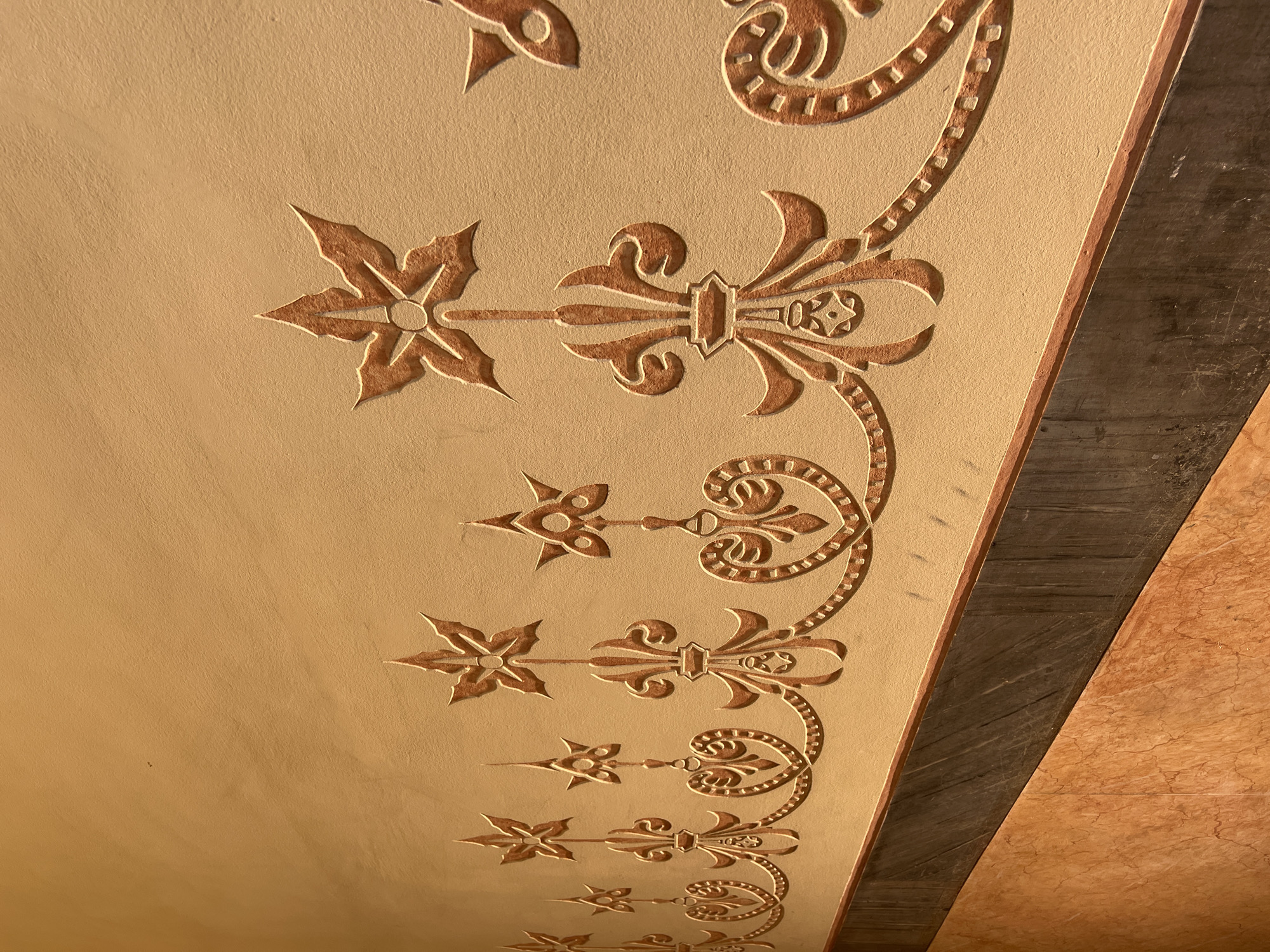The image shows a section of a decorative ceiling or wall in Barcelona, Spain. The design features a repetitive motif composed of a star-like element connected by a narrow line to a stylized fleur-de-lis-like shape. A curved, beaded element extends from the fleur-de-lis design, creating a visually continuous pattern. The decorative elements are a reddish-brown color, standing out against the cream or beige background of the ceiling/wall surface. The top right corner shows a transition to a different material, possibly a wooden or stone beam, that is brown in color and appears aged or weathered.