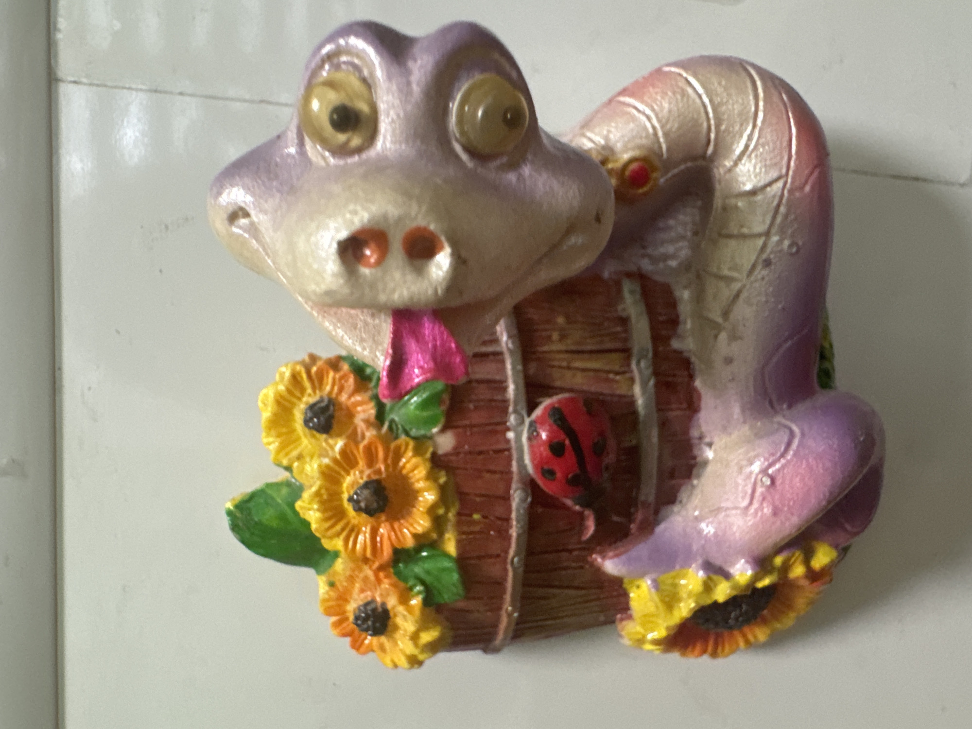 A whimsical, colorful sculpture of a creature resembling a dragon or serpent is depicted. The creature has a pale pink and purple body with iridescent highlights. Its head features large, round, yellow eyes with darker pupils and two orange spots on its snout. A bright pink ribbon is draped around its neck. The creature is curled around a wooden bucket filled with vibrant yellow and orange sunflowers, complete with green leaves. A small, red ladybug with black spots is perched on the side of the bucket. The sculpture is set against a plain, light-colored background, suggesting it may be a decorative item or a garden ornament. The scene is well-lit, with no apparent shadows indicating the direction of the light source. The overall impression is one of playful fantasy and nature.
