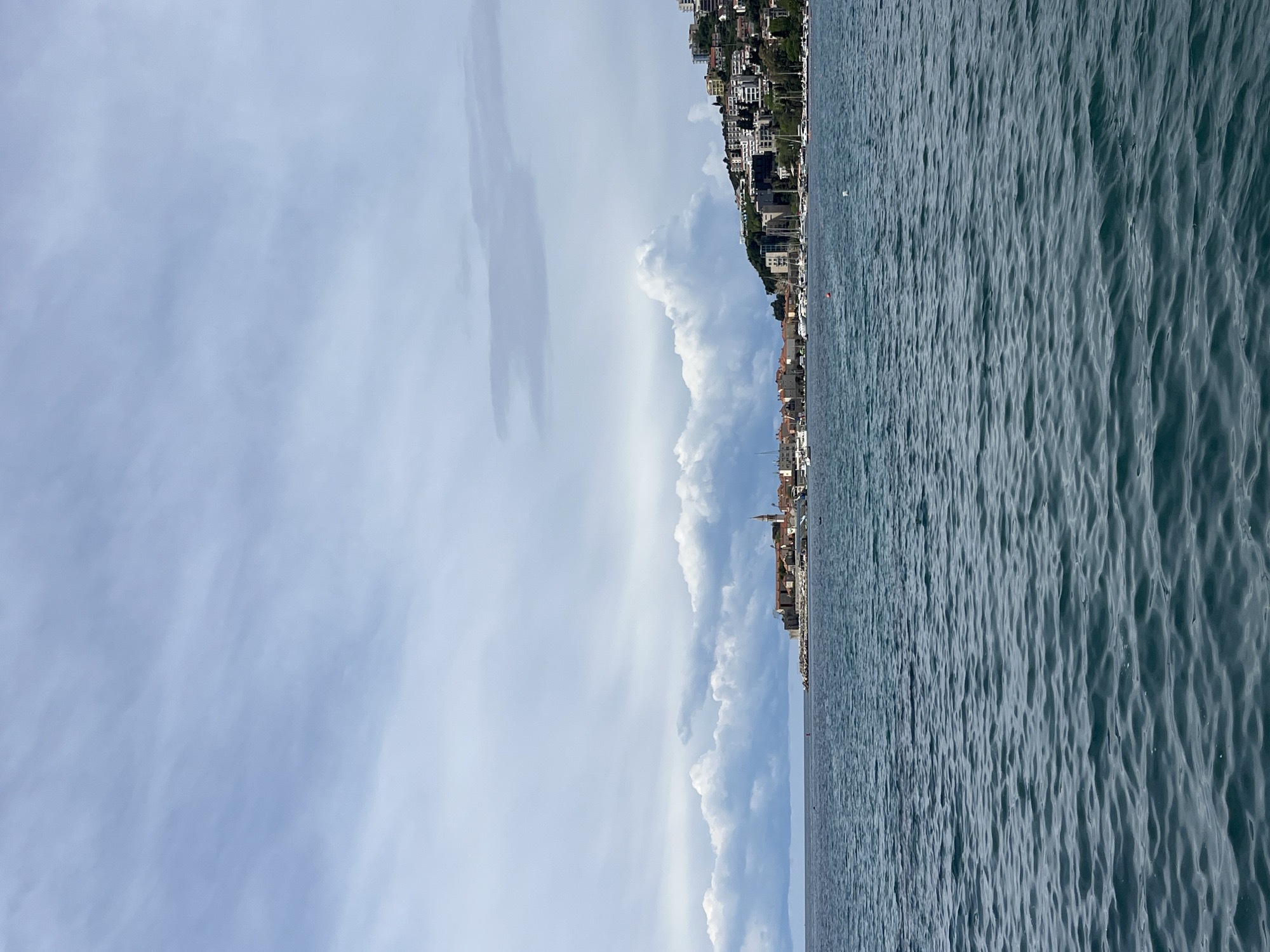The image, when viewed in its correct landscape orientation, displays a panoramic coastal scene of Budva, Montenegro. The horizon line runs horizontally across the middle of the frame, separating the sky and clouds above from the sea and coastline below.
The upper portion of the image is dominated by a light blue sky with scattered white and grey clouds. Towards the center-right, larger, more prominent cumulus cloud formations are visible, adding depth to the sky.
The coastline stretches across the mid-ground, featuring a densely built town or city situated along a sloping green hillside. Numerous buildings of varying architectural styles, many light-colored, can be seen ascending the terrain. Interspersed among the structures are pockets of lush greenery and trees. Hints of traditional red-tiled roofs and possibly church spires are visible within the urban sprawl.
The lower half of the image is occupied by a vast expanse of water, appearing as a deep blue-grey hue. The water's surface is textured with distinct, regular ripples, suggesting gentle movement rather than a perfectly smooth calm. Along the shoreline and extending into the water, several small boats or yachts are visible, some moored, others potentially anchored. Two small, reddish-orange buoys are also discernible in the water.
The scene depicts a clear, bright daytime, likely under partly cloudy conditions. No specific human activity or event is explicitly captured, presenting a tranquil and scenic view of the coastal area. There is no visible text in the image.