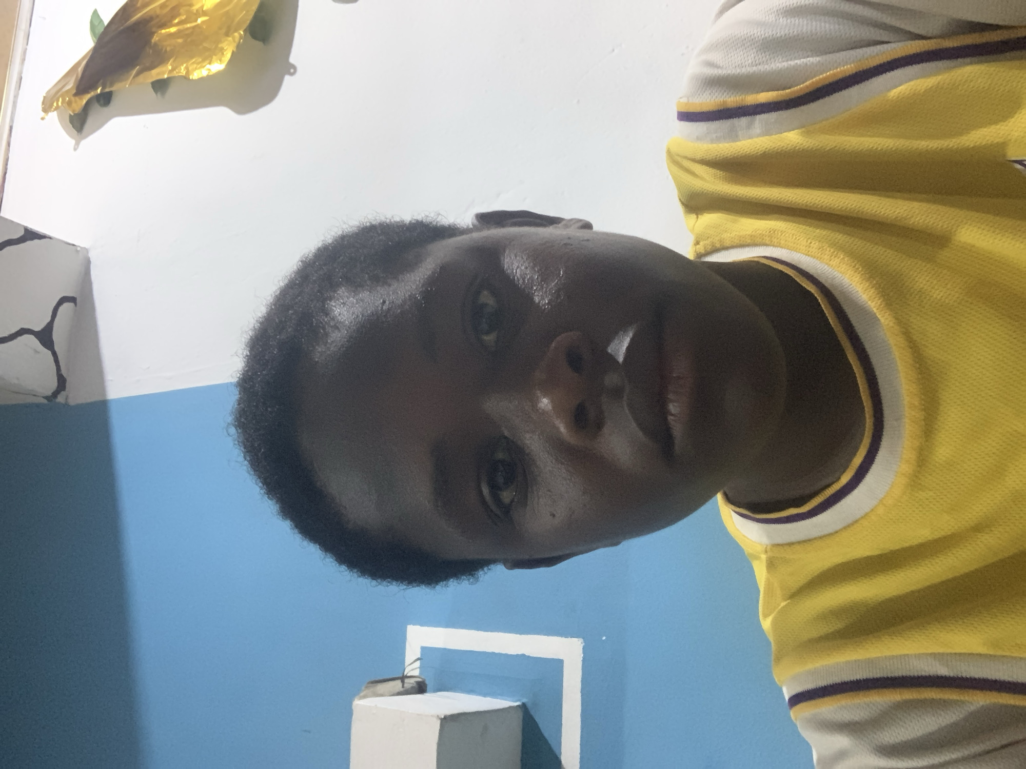 A person with dark skin and short, dark hair is wearing a yellow and white sports jersey with purple trim. The person's face is centered in the frame, with a slightly downward gaze. The background includes a white wall, a blue wall, and a white cube object. A gold foil decoration is visible in the upper left corner. The location is likely indoors, and cannot be confirmed to be in Jos, Nigeria.