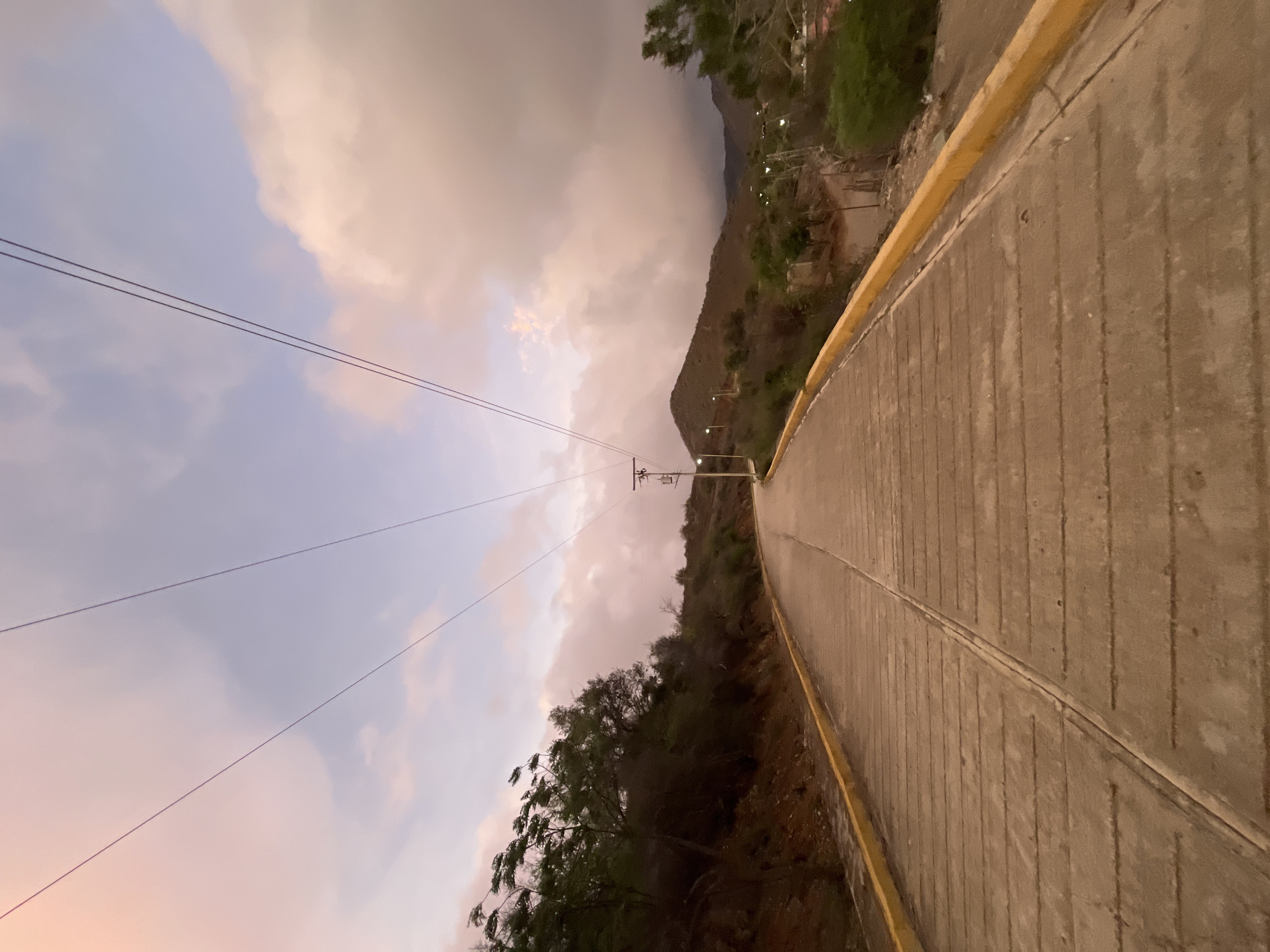 The image depicts a concrete road with a yellow curb, ascending a gentle incline. The road is flanked by arid vegetation and hills. Overhead, power lines crisscross the sky, which is filled with soft, diffused clouds tinged with pink and purple hues, suggesting either dawn or dusk. The scene is devoid of any people or vehicles, creating a sense of stillness. The landscape appears undeveloped, with a few scattered structures visible in the distance. The overall atmosphere is serene and natural.