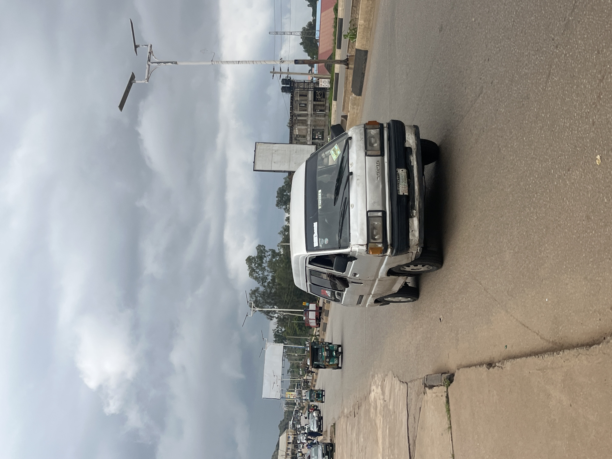 The image depicts a street scene in Gura Top, Nigeria, under a cloudy sky. A silver Toyota minvan is the primary subject, driving on the right side of a paved road. The minivan appears to be in use, with its front bumper slightly discolored and a license plate visible. Other vehicles, including several tricycles (popularly known as Keke Napep), cars, and a red truck, are also present on the road. The street is lined with a concrete sidewalk on the left and a raised concrete barrier on the right. In the background, buildings, trees, and streetlights can be seen. The overall environment suggests an urban or semi-urban setting with active traffic.