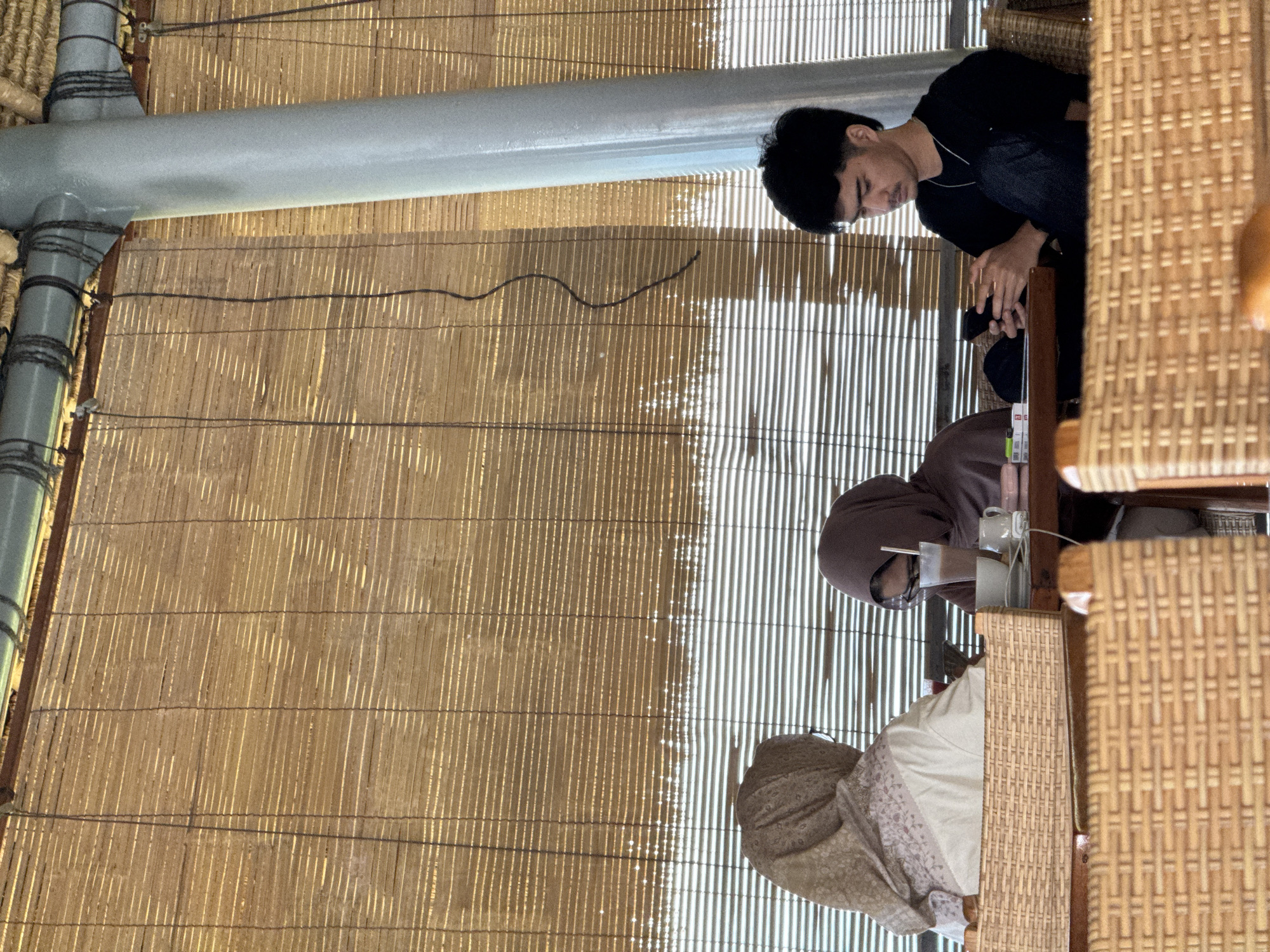 In Manggis, Indonesia, three individuals are seated at a table with woven bamboo blinds in the background. A woman wearing a light brown hijab and a white garment is on the left. In the center, another woman, also in a hijab but a darker brown, is looking down at a drink. To the right, a young man with dark hair and a black shirt is engrossed in his phone. The table is set with cups, a phone, and a small pink object, suggesting a casual cafe setting. The lighting is soft, indicating it could be daytime.
