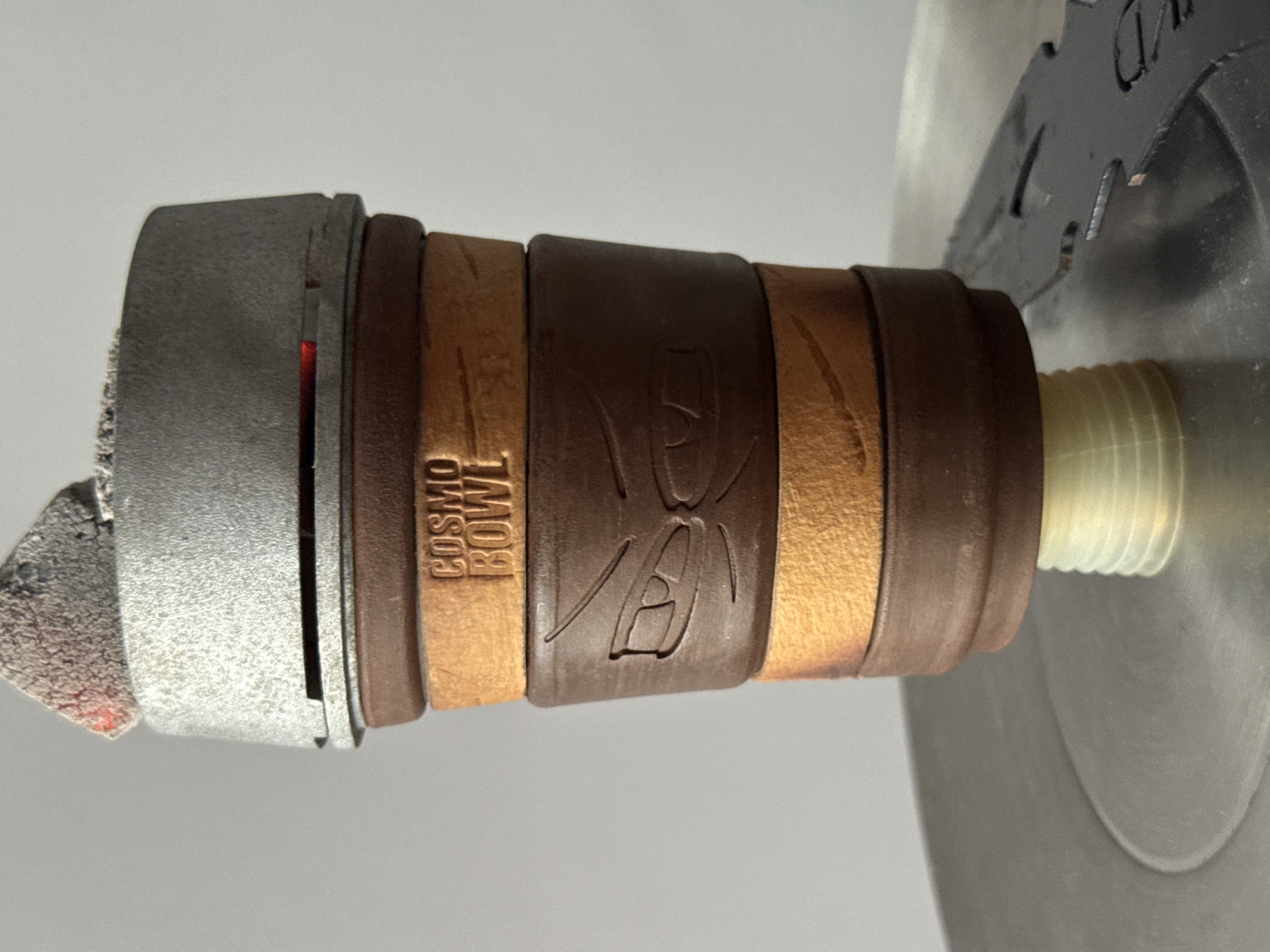 A close-up image shows a cylindrical object with alternating brown and light brown bands, partially affixed to a metal device with a threaded plastic component protruding from the right end. The band closest to the viewer bears an emblem, and a band labeled "COSMO BOWL" is visible. The object is indoors against a gray backdrop. The image doesn't provide enough context to pinpoint a specific location within Plovdiv, Bulgaria.