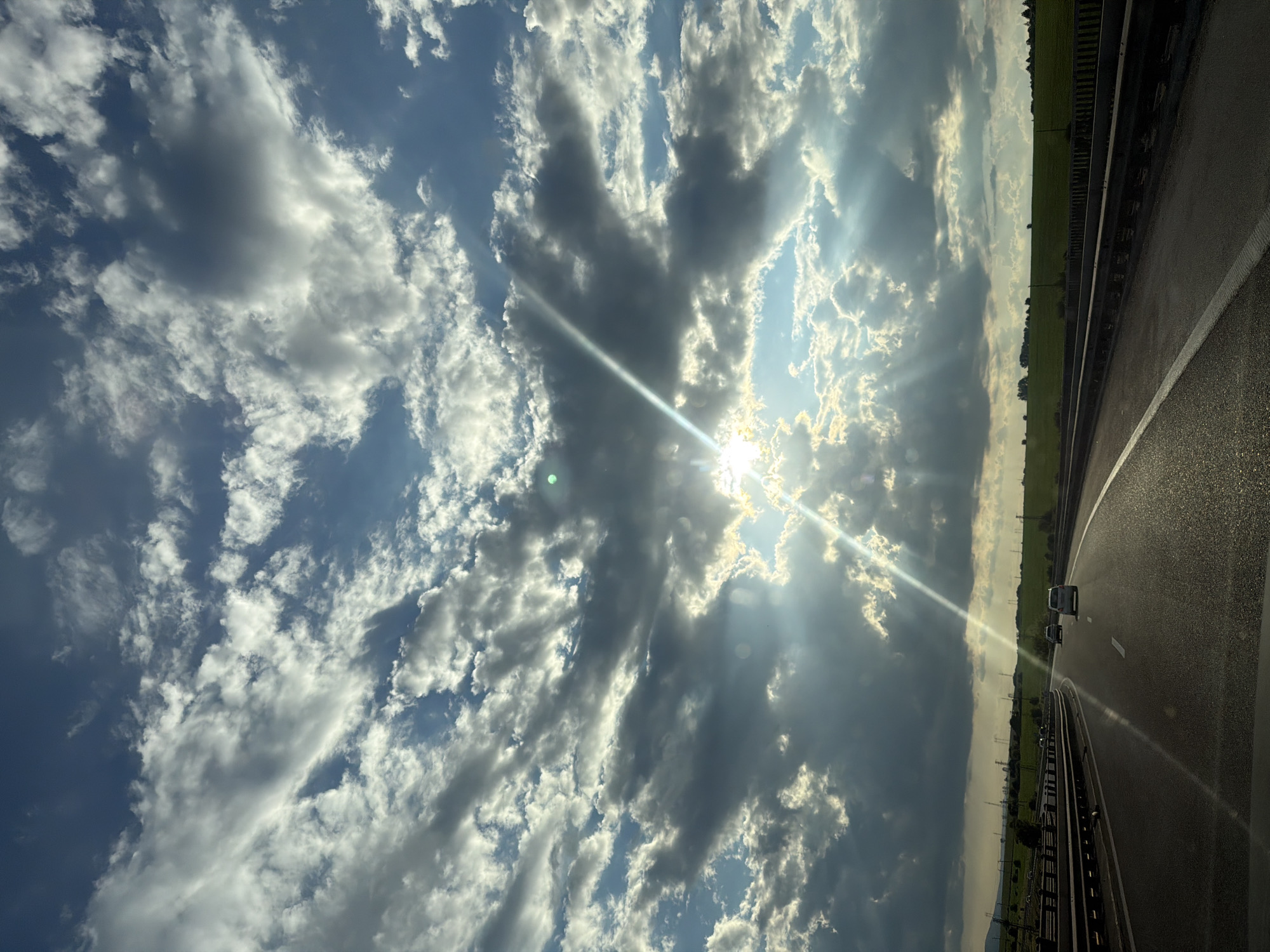 This image depicts a dynamic sky over a highway in Eleshnitsa, Bulgaria, viewed from the interior of a vehicle. The upper two-thirds of the frame are dominated by a sky filled with a mix of dense, dark gray cumulus clouds and brighter, scattered white clouds, against a backdrop of patches of clear blue sky. The sun, partially obscured by clouds, is positioned slightly to the right of the frame's vertical center, emitting strong, radiant crepuscular rays that extend downwards and across the sky. Multiple lens flares, including a prominent long streak originating from the sun and a smaller green circular flare on the left, are visible.
The lower third of the image shows a multi-lane asphalt highway curving gently to the right. The road surface is reflective, indicating either recent moisture or the direct glare of the sun. A metal guardrail runs along the right side of the highway. Three vehicles are visible ahead on the road: a dark-colored sedan in the left lane, and two white sedans further ahead in the right lane. No occupants are discernible.
The landscape flanking the highway consists of green fields and rolling hills under the dramatic sky. Scattered trees and shrubs are visible, particularly on the left side of the road. In the distance, numerous electrical transmission towers with power lines span across the horizon, suggesting an agricultural and industrial landscape. The lighting is characterized by high contrast, with bright sunlit areas and deep shadows cast by the clouds. No people are explicitly visible in the scene.