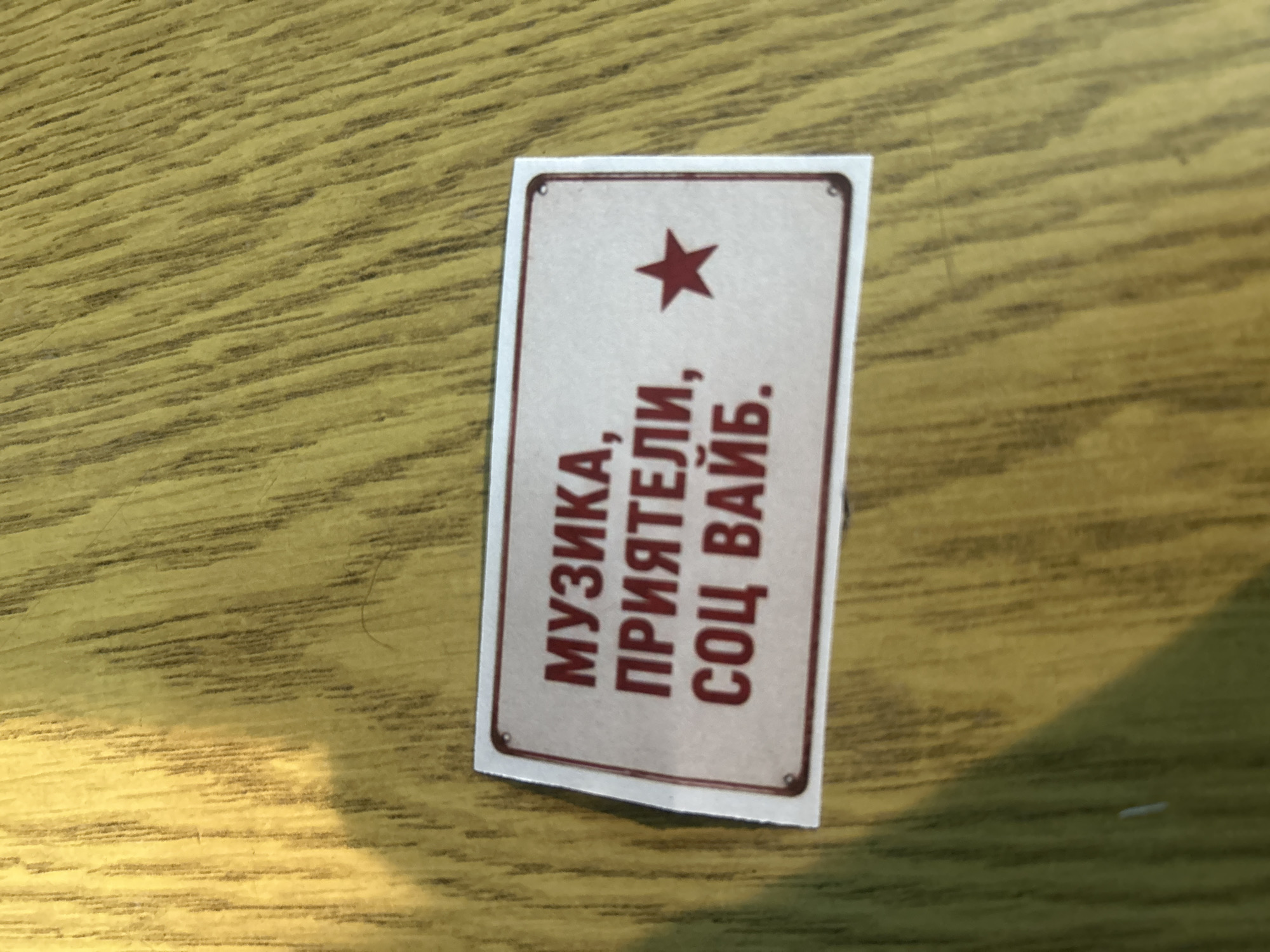A sticker or sign rests on a wood-grain surface. The sign is rectangular with rounded corners and a thin border. A five-pointed red star is at the top, above the text "МУЗИКА, ПРИЯТЕЛИ, СОЦ ВАЙБ." in red. The sign is slightly crooked. There is no indication of Veliko Tarnovo, Bulgaria.
