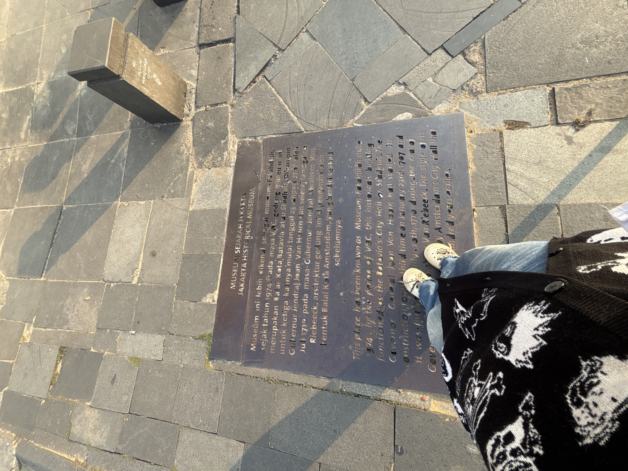 The image shows a close-up view of a plaque embedded in a stone pavement, with a person's legs and feet in the foreground. The plaque has text in both Indonesian and English. The Indonesian text reads: "MUSEUM SEJARAH JAKARTA. Museum ini lebih dikenal sekarang sebagai Museum Fatahillah, sejak tahun 1974. Pada masa VOC, gedung museum ini merupakan Balai Kota Batavia. Dibangun tanggal 23 Januari 1707 oleh Joan van Hoorn dan selesai tanggal 10 Juli 1710 pada masa Gubernur Jendral Abraham. Van Riebeeck, arsitektur gedung ini dipengaruhi oleh bentuk Balai Kota Amsterdam, yang berdiri sembilan abad sebelumnya."
The English text reads: "This place has been known as Museum Fatahillah since 1974. In the period of VOC, this historical building functioned as the Batavia's City Hall. The construction of Governor General Joan Van Hoorn had started its third time on January 23rd, 1707 and it was finished on July 10th, 1710 during the era of Van Riebeeck. The style of its by Amsterdam's City Hall that hundered years earlier."
The person is wearing blue jeans and white sneakers with a black and white patterned cardigan. The pavement is made of square and rectangular stone slabs. The overall scene suggests an outdoor location, likely a historical site in West Jakarta, Indonesia, given the text on the plaque. The sunlight casts shadows on the pavement, indicating it is daytime.