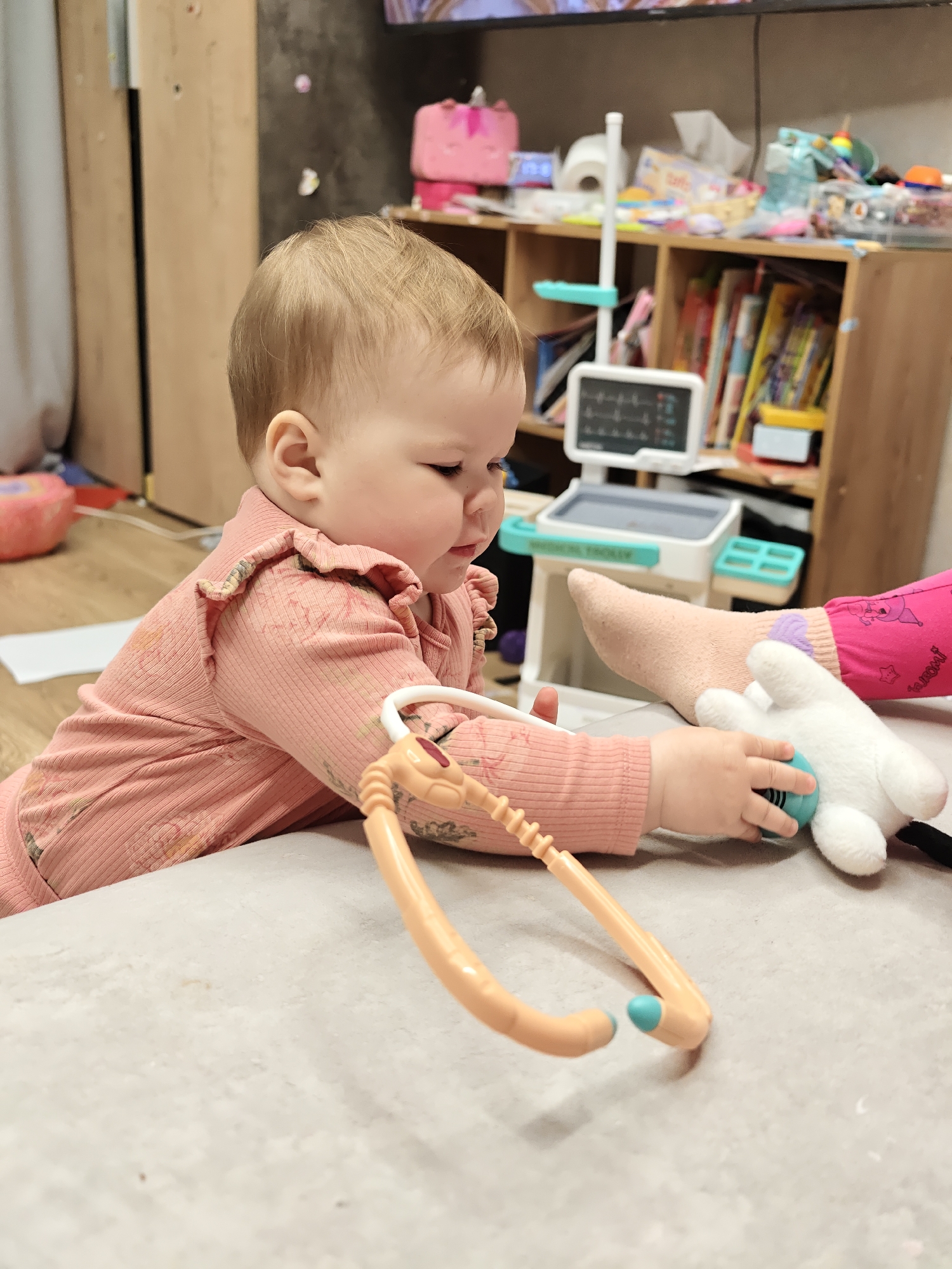 A baby with fair hair, wearing a pink ribbed long-sleeved top with ruffled shoulders, is shown in a close-up, indoor shot. The baby is focused on playing, holding a toy stethoscope in their right hand. The light orange stethoscope has blue ear tips and a blue-green diaphragm, which the baby is pressing against a white stuffed animal. Another person's leg and foot, covered by a light pink sock and part of pink trousers, are visible next to the stuffed animal, suggesting the baby is "examining" it.
In the background, a child's toy medical trolley is visible, with "MEDICAL TROLLEY" printed on its side. It features a screen displaying medical readings and various compartments. Behind the trolley, there are wooden shelves filled with numerous books, toys, and miscellaneous household items, including a roll of toilet paper and tissue boxes. A television screen is partially visible at the top of the frame. The setting appears to be a cozy, domestic space, possibly a living room or play area, with a light-colored surface (likely a couch or play mat) in the foreground where the baby is situated. The overall atmosphere is one of innocent play and exploration. The baby's expression is one of concentration, indicating deep engagement with their toy.