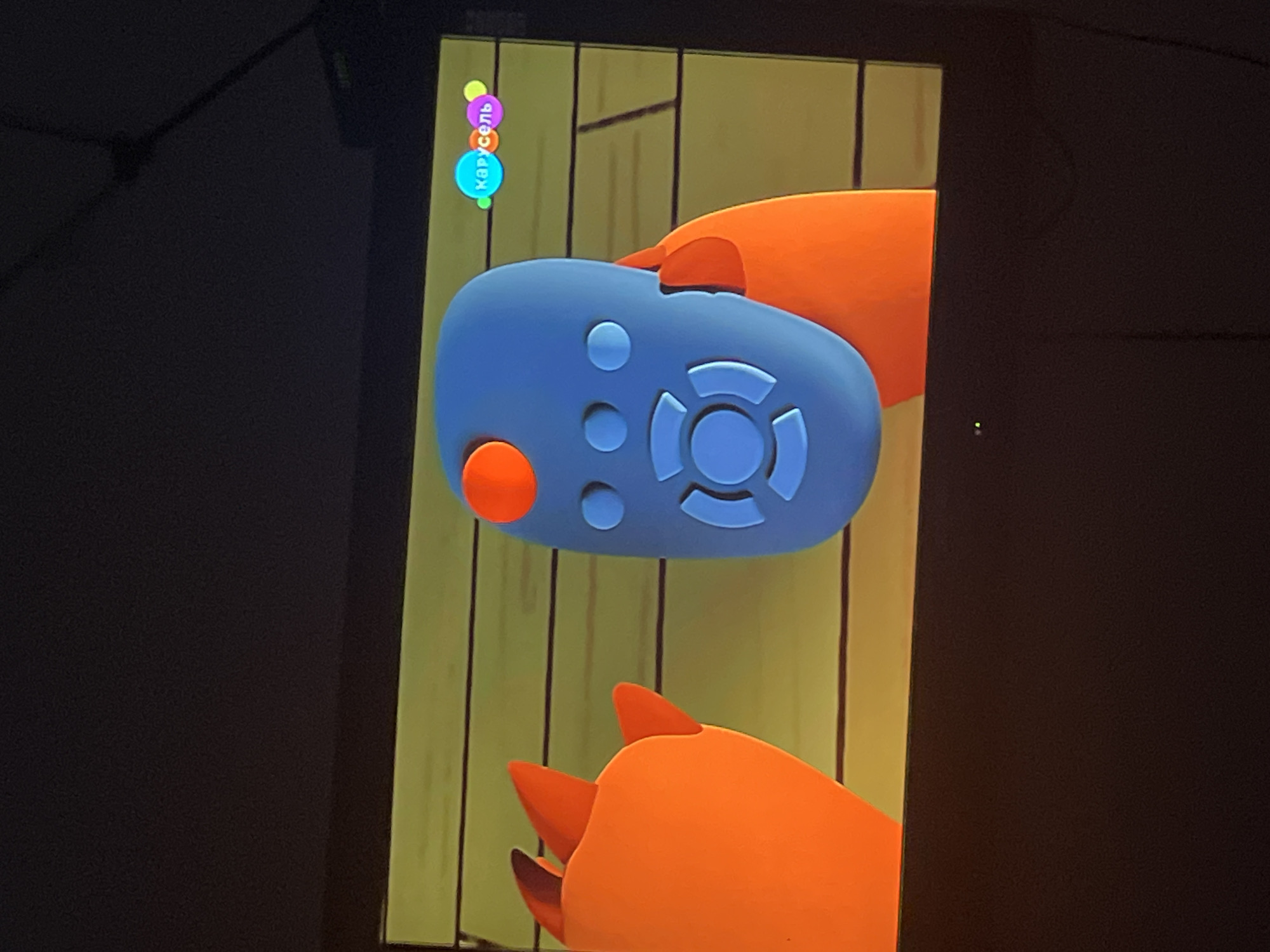 The image shows a cartoon television program displayed on a screen. The program features two animated characters and a remote control. On the left, an orange cartoon character with pointed ears is visible. In the center, a blue cartoon remote control with orange and light blue buttons is being held by another orange character's appendage. To the right of the remote, a logo with colorful circles and the word "Карусель" (Carousel) in Russian is displayed. The background of the program appears to be a wooden wall. The image is a photograph of the television screen, with the room around the screen being dark. There is no information in the image that allows for the determination of its location in Chelyabinsk, Russia.