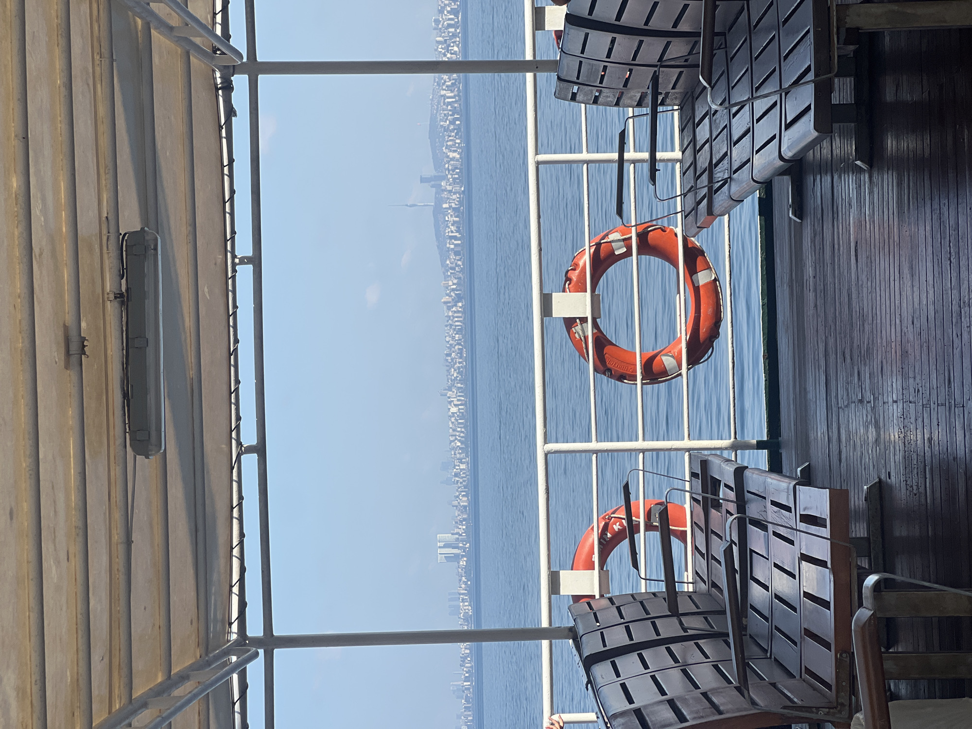 A wide shot captures an outdoor seating area of a ferry with a view of the Istanbul skyline. The foreground features two rows of empty wooden benches with metal frames. The benches are positioned on a dark, wet wooden deck. In the mid-ground, a white metal railing separates the deck from the water. Two bright orange life preservers are hung on the railing, one on the left and one on the right. Beyond the railing, the calm blue sea stretches towards a distant city skyline. The skyline is characterized by numerous buildings of varying heights, with a prominent tower visible in the center. The sky is a clear, light blue. The image is taken from the perspective of someone seated on the ferry, looking out towards the sea and the city. The scene suggests a calm day on the water, likely a passenger ferry route connecting to or from the Prince Islands, Turkey.