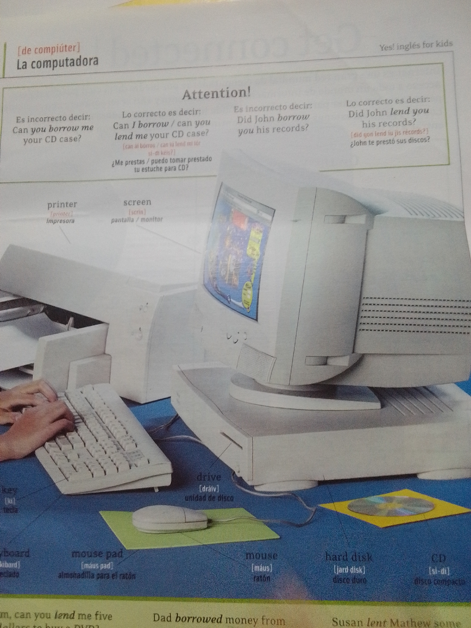This is a close-up, eye-level, indoor photograph of a page from a children's English language learning book. The page features illustrations and text related to computers and vocabulary. The main focus is on a vintage desktop computer setup, including a monitor, a tower, a keyboard, and a mouse on a mousepad. There is also a printer to the left of the computer. Labels in English and Spanish identify various computer components such as "printer," "screen," "keyboard," "mouse," "mouse pad," "drive," "hard disk," and "CD."
The page also includes example sentences and grammar corrections, with headings like "Attention!" and phrases such as "Es incorrecto decir" (It is incorrect to say) and "Lo correcto es decir" (The correct thing to say). The bottom of the page contains additional sentences related to borrowing and lending, with handwritten annotations in some areas. The overall lighting is bright, suggesting an indoor environment with good illumination. There is no visible indication of La Tagua, Venezuela, or any outdoor elements in the image.