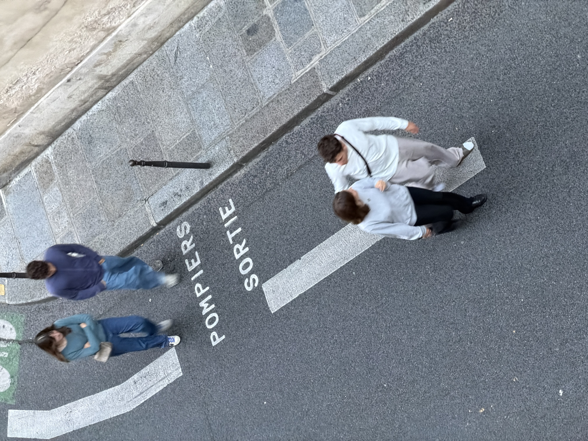 The image, taken from an elevated viewpoint, shows a section of a street in Paris, France. The street surface is asphalt, and there is a painted designation of "POMPIERS SORTIE" on the road, indicating an exit for firefighters. Four people are visible on the street. Two are walking together, one wearing a white top and light-colored pants, and the other wearing a light blue top and dark pants. The other two are also walking together, one wearing a blue sweater and the other wearing a blue sweater and blue jeans. The sidewalk is made of stone blocks. A short, black pole is near the sidewalk's edge. The angle of the shot suggests it was taken from a building overlooking the street.