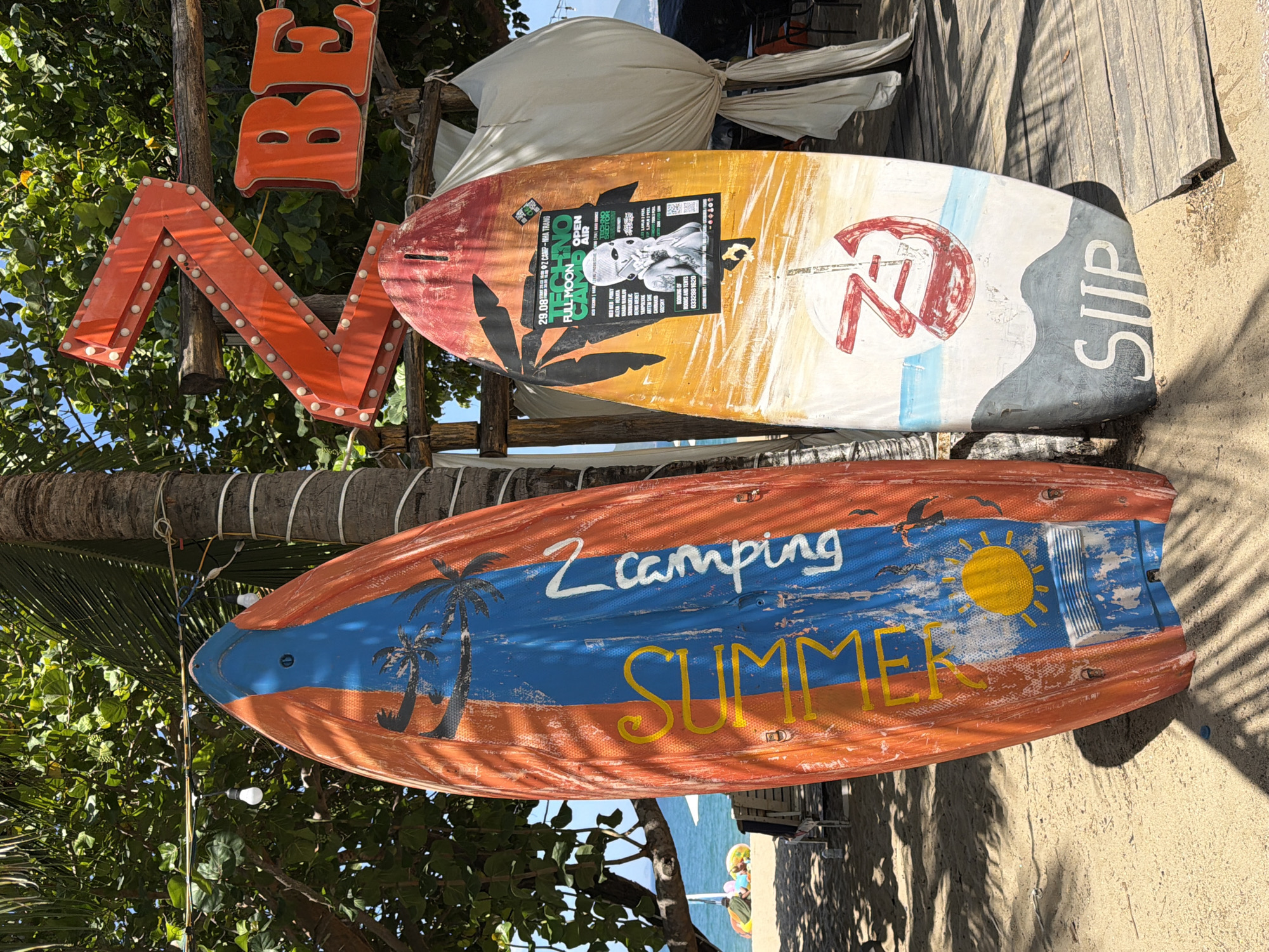 Two surfboards are propped upright on a sandy beach, with a palm tree and lush green foliage in the background. The surfboard on the left is primarily orange with a blue stripe down the middle and features a depiction of palm trees and the word "SUMMER" in yellow lettering. The surfboard on the right is white with a colorful graphic, a flyer for "TECHNO FULL MOON CAMP OPEN AIR," and the word "SUP" at the bottom. To the right of the surfboards, a large, orange, illuminated "Z" sign is visible, followed by an orange "BE" sign. The ocean and a section of beach with lounge chairs and umbrellas can be seen in the distance. The overall scene suggests a beachside location in Nha Trang, Vietnam, likely a resort or a popular spot for water sports and events.