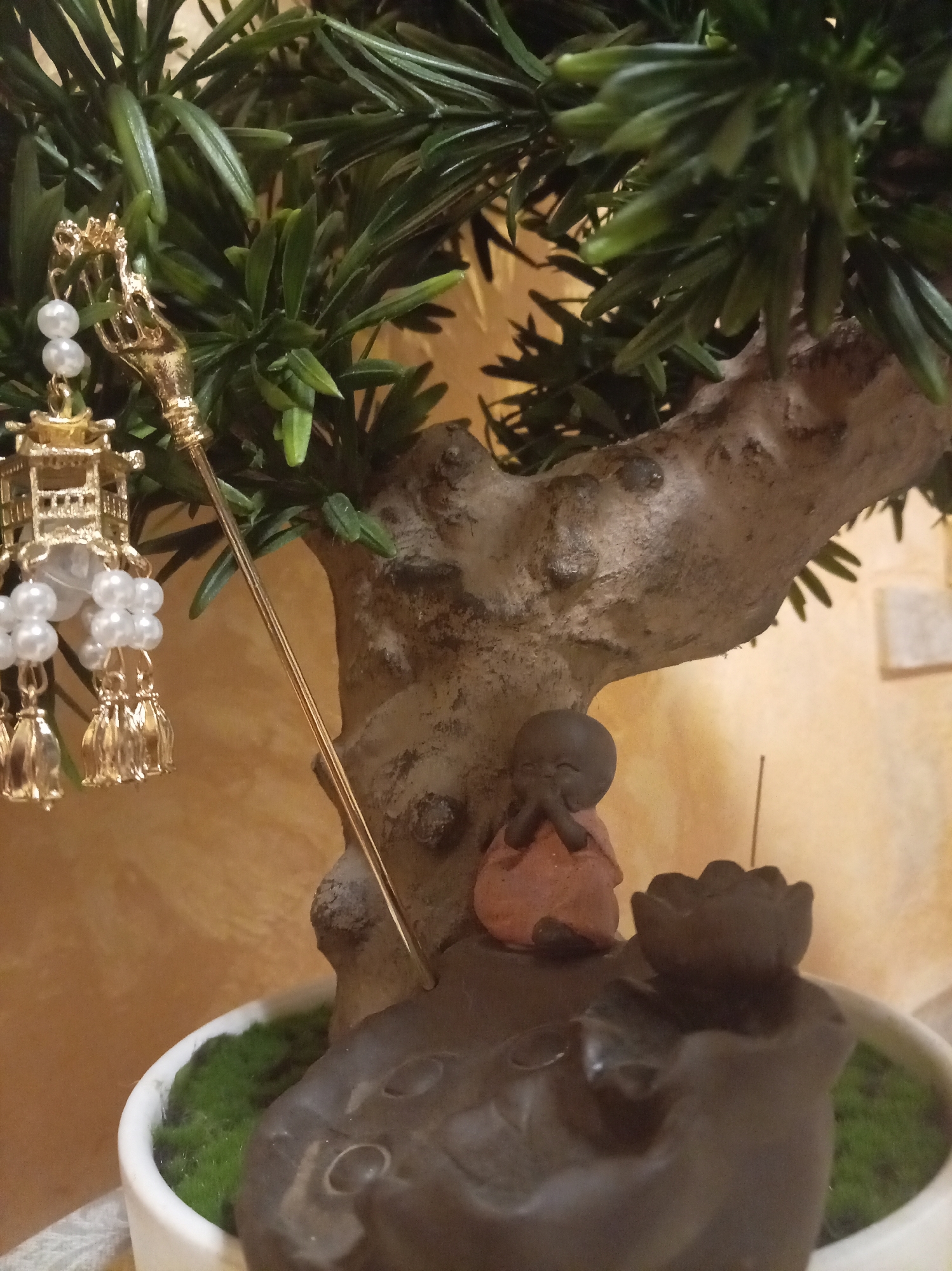 A close-up shot shows a decorative item resembling a miniature bonsai tree, adorned with a gold-colored ornament featuring a pagoda, pearls, and tassels. Beside the tree trunk is a small figurine of a monk in an orange robe, with hands clasped in prayer. To the right of the monk is a dark brown, lotus-shaped incense holder with a thin stick inserted, emitting a faint wisp of smoke. The scene is set against a textured, ochre-colored background, with glimpses of green moss and a white pot at the base of the tree. The lighting is soft, highlighting the details of the objects.