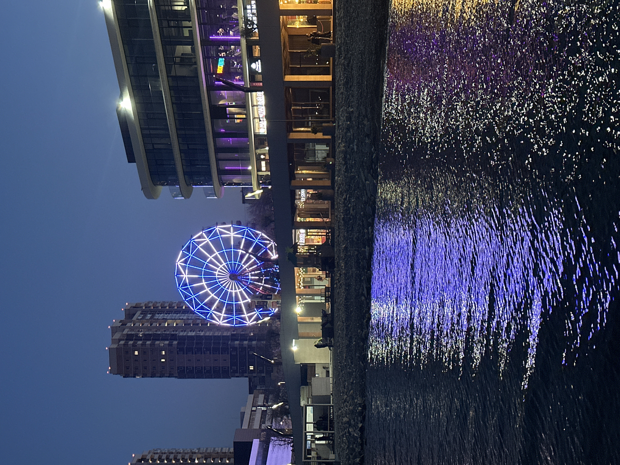 This night-time image, set in Novorossiysk, Russia, captures a lively waterfront scene. On the left, a series of modern, multi-story buildings feature extensive glass facades and warm interior lighting, alongside vibrant exterior neon lights in purple and yellow hues, suggesting shops and restaurants.
Dominating the mid-ground is a large Ferris wheel, brilliantly illuminated with intricate patterns of blue and white lights, standing out against the dark sky. Below and behind it, other urban structures are faintly visible.
To the right, a body of water serves as a dynamic canvas, reflecting the bright lights of the buildings and the Ferris wheel. The rippled surface of the water transforms these reflections into shimmering, elongated streaks and sparkling dots of blue, purple, and white light. A dark, textured wall separates the land from the illuminated water.
A few indistinct figures of people can be seen near the buildings, suggesting evening leisure activity. The scene conveys a vibrant, urban atmosphere. Visible text on some building signs is partially obscured but includes what appears to be "DREAMLAND" and potentially "COFFEE" on different establishments.