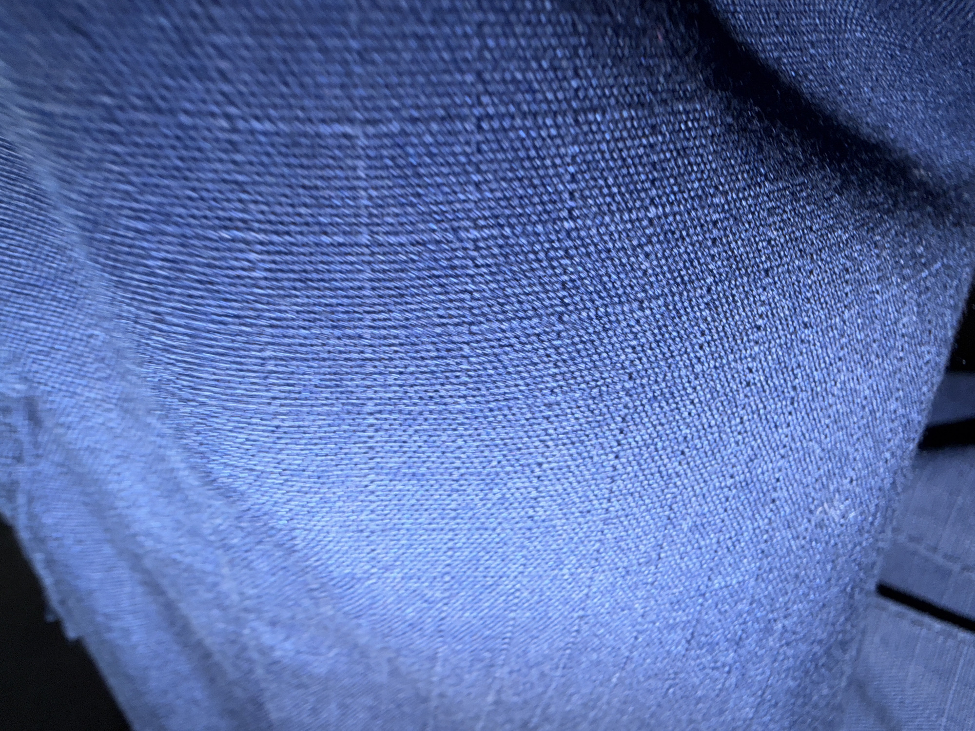 The image displays a close-up, abstract view of dark blue textured fabric, possibly denim or a similar material. The texture shows a distinct woven pattern with raised threads creating a ribbed effect. The lighting creates highlights and shadows across the fabric, emphasizing its three-dimensional qualities. The bottom of the image reveals a glimpse of black material with a linear pattern, suggesting it might be a seam or another part of the garment. There is no discernible urban environment or any indicators that this image is located in Shingak-Kul', Russia. The scene is entirely focused on the fabric's detail.