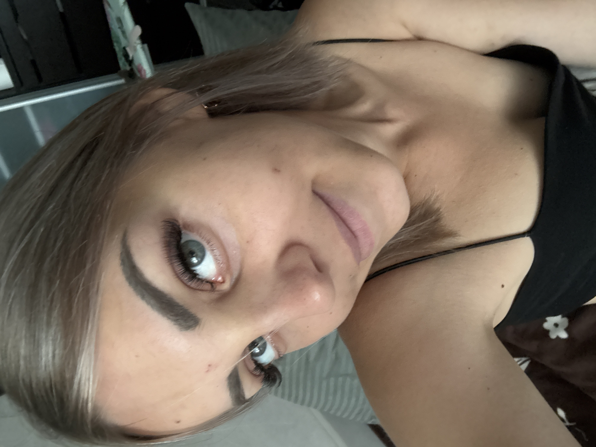 A young Caucasian woman with fair skin, blonde hair, and blue eyes takes a selfie. She is wearing a black camisole and has a subtle smile. Her makeup includes false eyelashes and well-defined eyebrows. The background is indistinct, with hints of bedding and furniture, suggesting an indoor setting. The lighting is soft, and the focus is on her face.