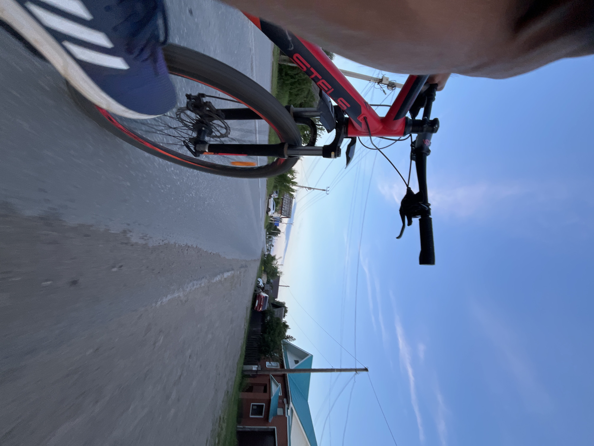 The image depicts a first-person perspective of someone riding a red and black bicycle on a paved road. The bicycle's front wheel, handlebars, and part of the frame are visible in the foreground. A person's leg wearing a blue athletic shoe with white stripes is also in the lower left corner. The road appears to be a country road or a road in a rural settlement, with a dirt shoulder on the right side. Along the right side of the road are several houses with red brick and turquoise roofs, surrounded by fences and trees. Utility poles and power lines are visible along the road. The sky is blue with some wispy clouds, suggesting it is late afternoon or early evening. The scene is likely located in a rural area of Chaikovsky, Russia, given the architectural style of the houses and the overall environment.