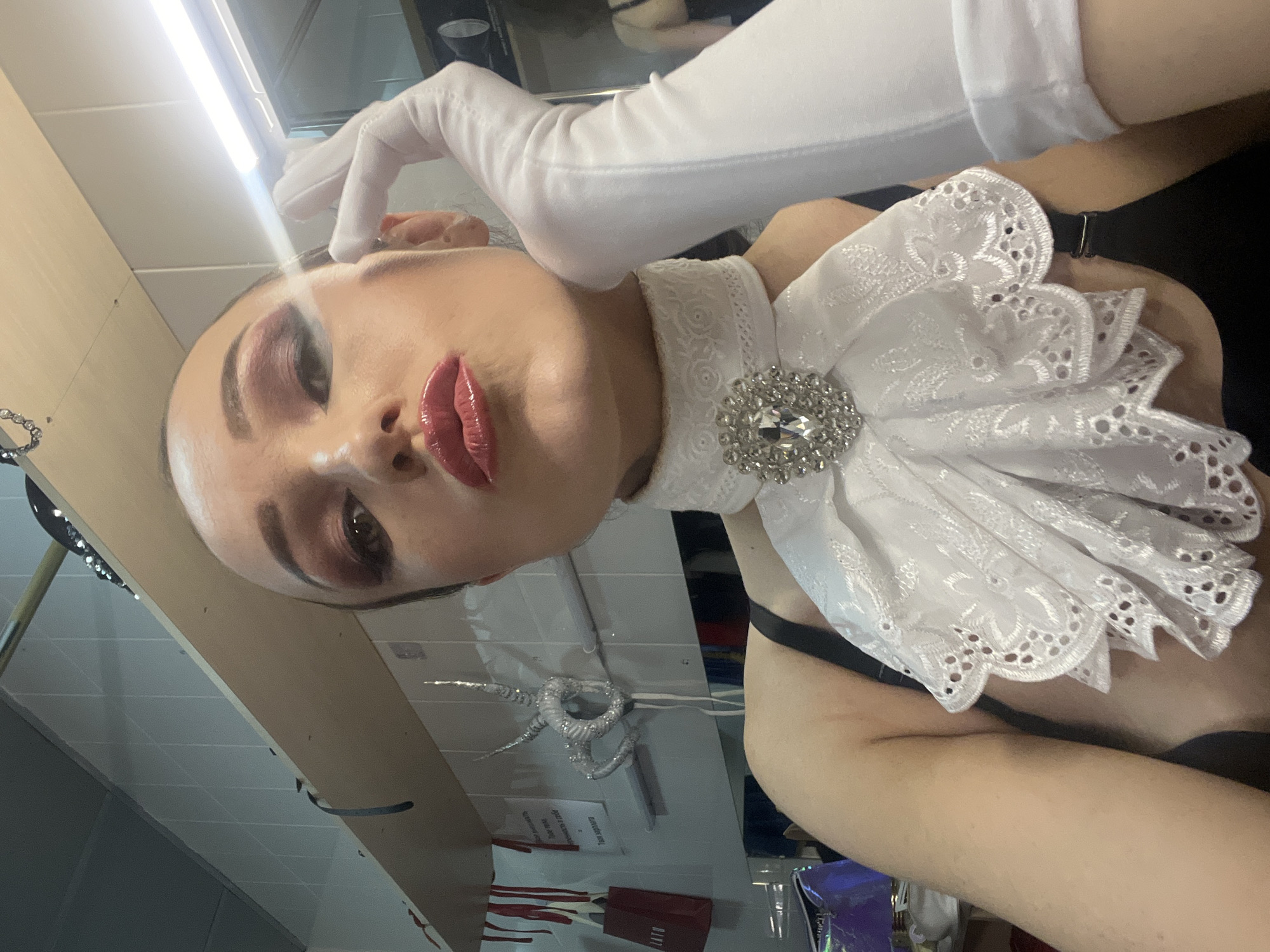 A person with dark hair and full makeup, wearing a white jabot with a jeweled brooch, white gloves, and a black top, poses for a selfie in a backstage area. The background consists of white tiled walls, shelves with various items including a red bag, and a mirror. The person is looking at the camera with their hand touching their cheek, and their lips are pursed. The lighting is bright, originating from overhead fluorescent fixtures.