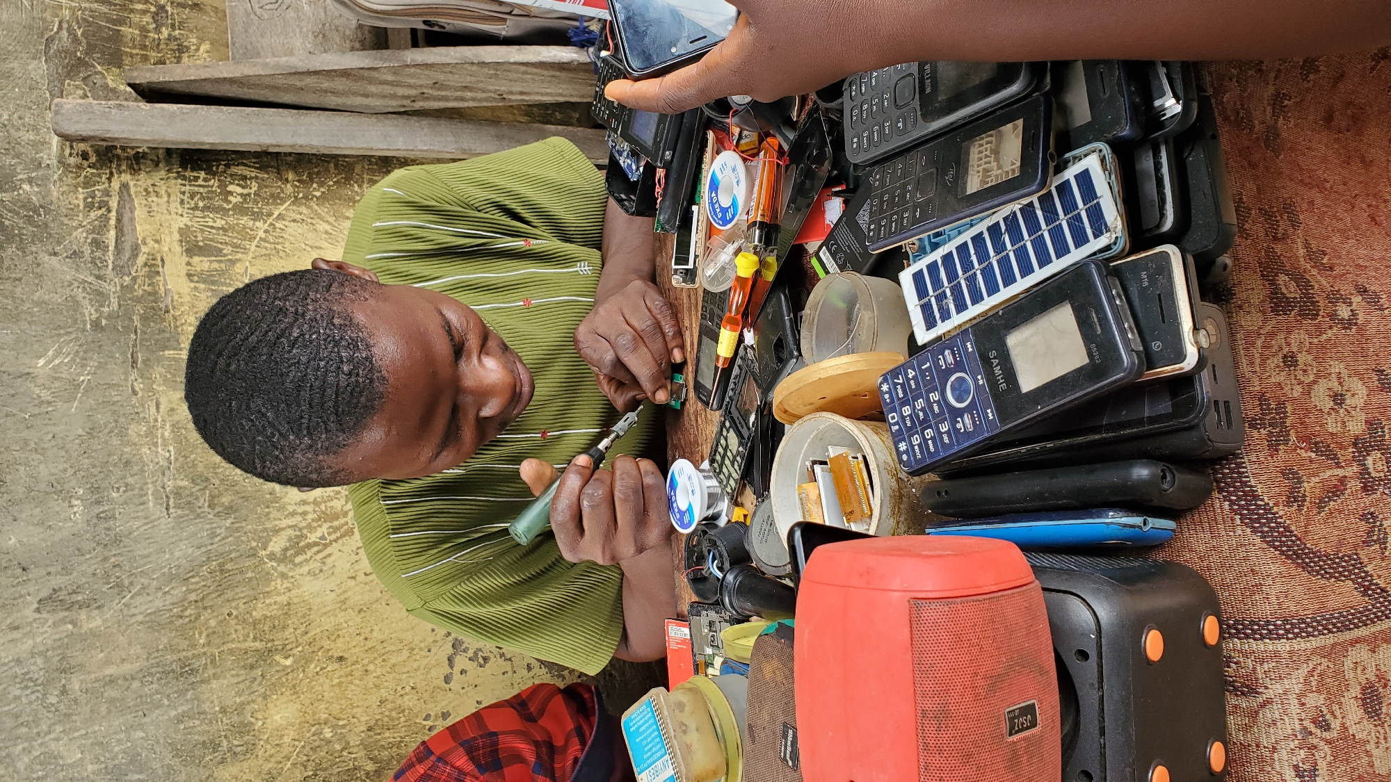 A young Nigerian man, wearing a green and white striped short-sleeved shirt, is focused on repairing a circuit board on a wooden table. He is holding a soldering iron and appears to be soldering a component. The table is covered with various electronic devices, including disassembled mobile phones, batteries, a red speaker, and a black power bank. In the background, a textured, weathered wall is visible, characteristic of an outdoor or semi-enclosed workspace in Jalingo, Nigeria. The overall scene depicts a busy, informal electronics repair shop.