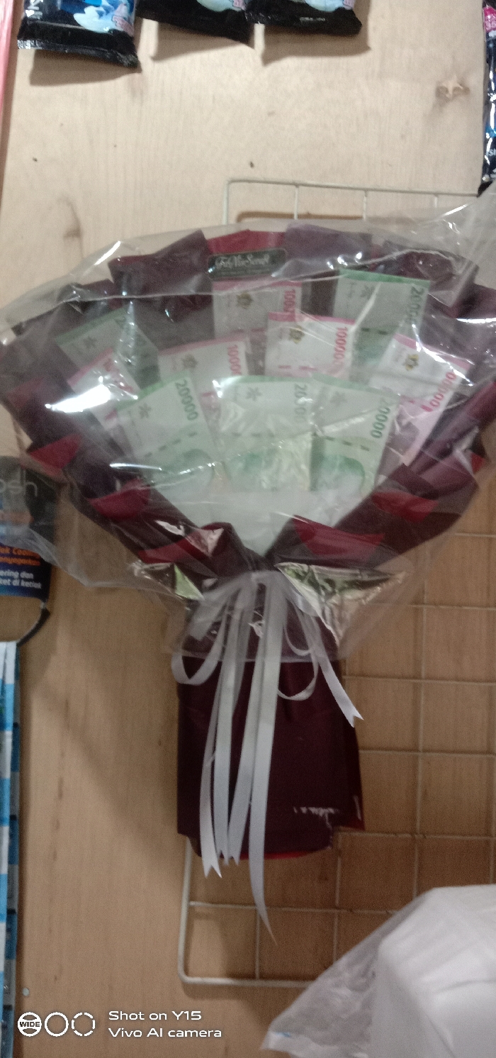 A money bouquet, wrapped in clear plastic and maroon paper/fabric, is the central subject, photographed indoors in Temenggungan, Indonesia. It is adorned with a white multi-loop ribbon tied at its base. The bouquet contains Indonesian Rupiah banknotes of visible denominations: 10,000 (green), 20,000 (green), and 100,000 (pink), arranged to form the 'flowers.' A small dark label, partially obscured, is visible through the plastic wrap near the top, possibly reading 'Fir Sense.' The background on the left features a light-colored wooden wall from which dark-colored packages are suspended. A blue and white patterned fabric and a sign with Indonesian text are also partially visible on the left. The background on the right is composed of light brown square wall tiles, with a white wire grid shelf mounted below the bouquet. No individuals or active interactions are depicted.