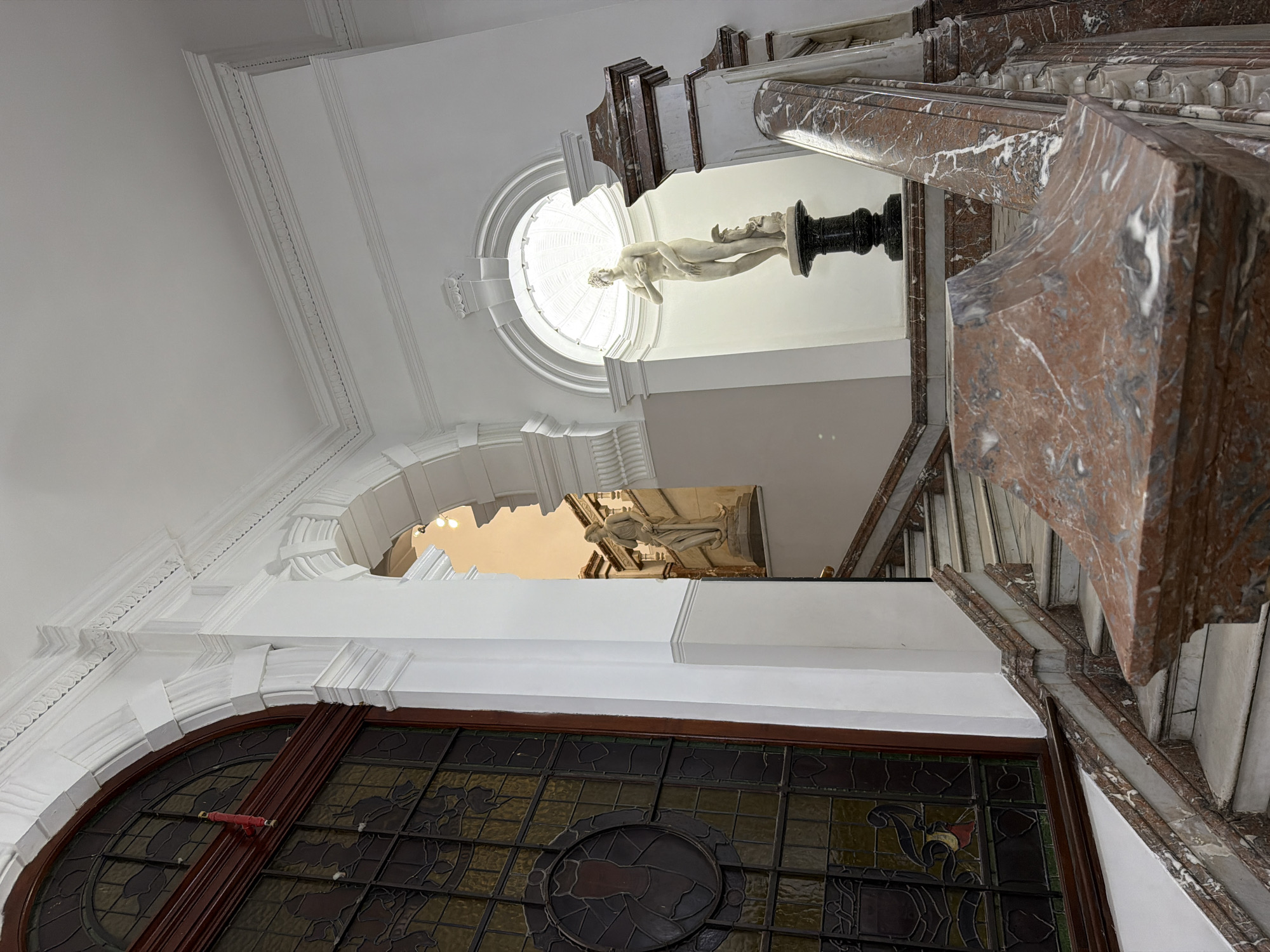 The image depicts the interior of the Iziko South African National Gallery in Cape Town, South Africa. It focuses on the architectural details and decorative elements within the building. A large arched window with intricate stained glass patterns dominates the foreground. Above the window, the architecture transitions into a white, ornate archway. Further up, the image captures a staircase with marble steps and a handrail featuring distinctive reddish-brown marble patterns. At the top of the staircase, there is a statue, seemingly elevated on a pedestal. A circular window or skylight provides natural light, illuminating the statue and surrounding area. The ceiling is white with decorative moldings.