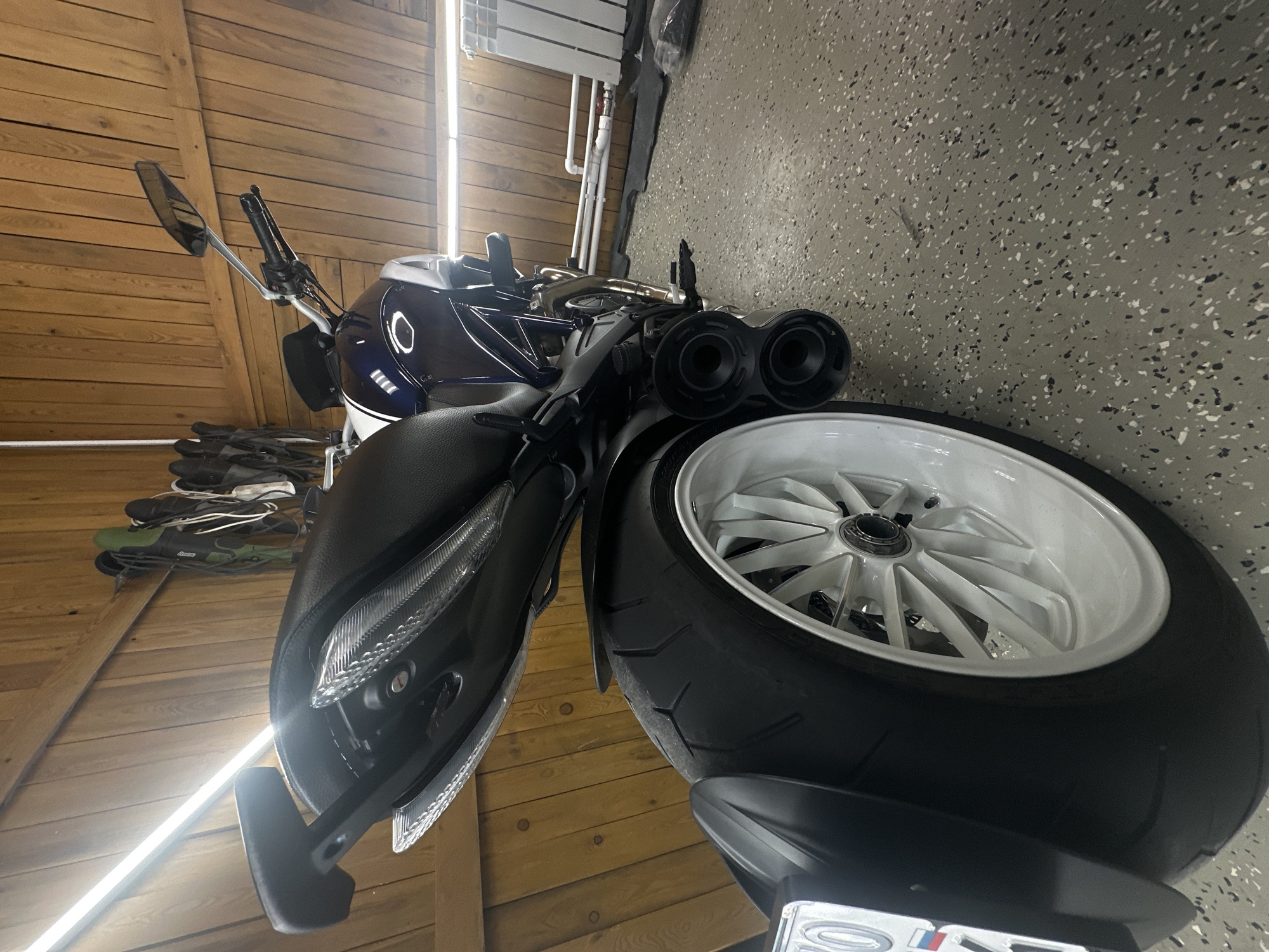 The image shows a motorcycle parked in what appears to be a garage. The motorcycle is dark blue and white with a black seat. It has a white rear wheel. The garage has wooden walls on one side and a gray, speckled floor on the other. There are shoes and other items near the wooden wall. An overhead light is visible along the wooden wall. The location is Unknown, Unknown. There are no people visible.