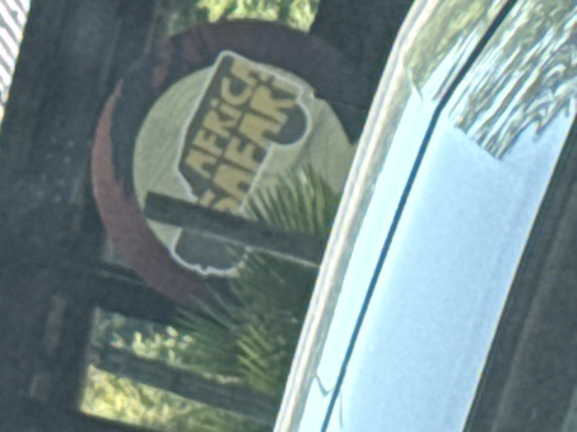 The image features a partially obscured sign that reads "Africa Safari." The sign has a circular design with brown and green colors. The sign is partially blocked by greenery and the frame of what appears to be a window. The location is likely near or at the Africa Safari Park in Bussolengo, Italy.