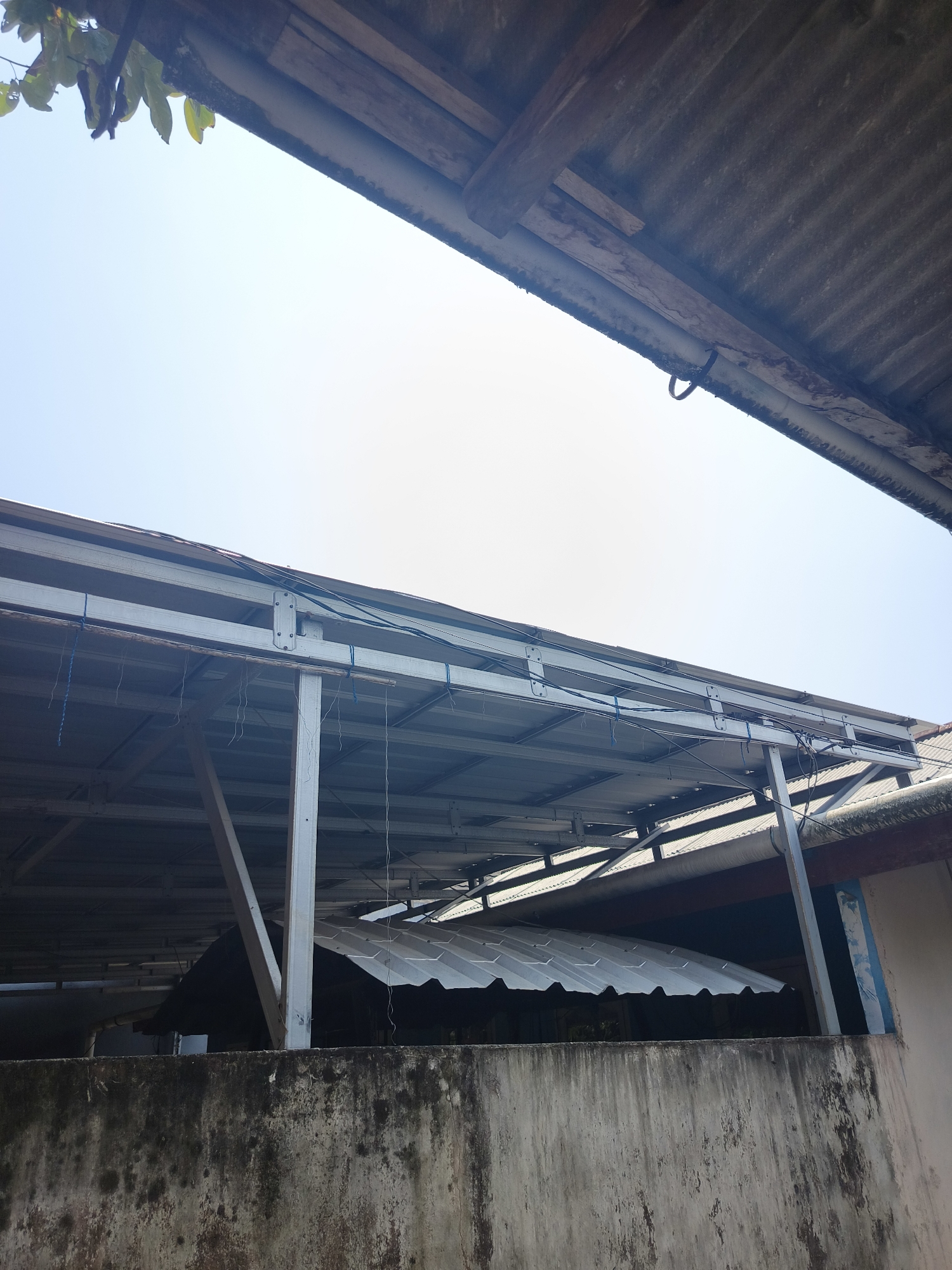 The image shows a view of rooftops and a clear sky in Pangkalpinang, Indonesia. In the foreground, a weathered concrete wall with moss and stains is visible. Above the wall, a light-colored metal structure with a corrugated metal roof is supported by metal beams. There are also some blue and white strings hanging from the structure. In the upper portion of the image, the edge of another corrugated metal roof is visible, along with some wooden elements and leaves from a tree. The sky is bright and clear, indicating daytime. The scene appears to be an outdoor area, possibly a backyard or a space between buildings.
