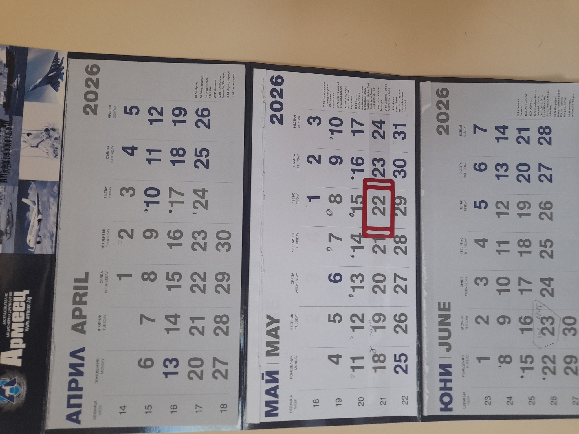 The image is a close-up shot of a Bulgarian calendar displaying three months: April, May, and June of the year 2026. The calendar is likely hanging on a wall. The month of May is prominently featured with a red rectangle highlighting the date 22. Each month's grid shows the weekdays in Bulgarian: Monday, Tuesday, Wednesday, Thursday, Friday, Saturday, Sunday. The top of the calendar features images of aircraft and the logo of an insurance company, "Армеец" (Armeec), with the website address www.armeec.bg. There are also descriptions for important Bulgarian holidays in each month. The calendar is likely located in Bulgaria, but not specifically in Sofia.