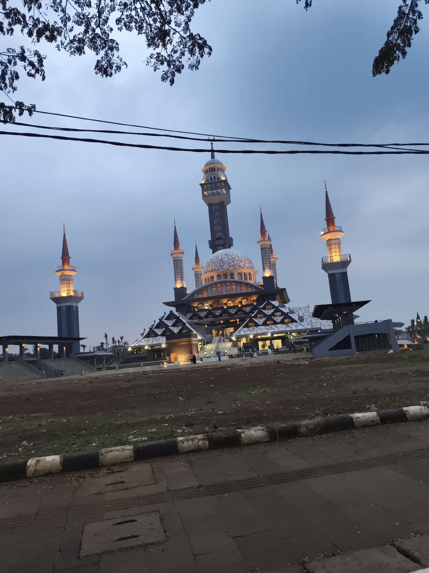 The image presents a grand, illuminated mosque under a twilight sky in Cibinong, Indonesia. The sky is a deep, muted blue-grey, suggesting dusk or early evening, and is partially framed at the top by dark, silhouetted tree branches. Two dark utility wires traverse horizontally across the upper portion of the scene.
The mosque is the dominant subject, showcasing intricate architecture with a prominent central dome and several slender minarets of varying heights. These minarets and key sections of the mosque are brightly lit with warm, yellow-orange artificial lights, emphasizing their design and the overall grandeur of the structure. The main dome is light-colored, possibly white or light grey, adorned with subtle patterns. The mosque's lower sections feature complex, multi-tiered rooflines and steps leading up to its main entrances.
Several people are visible around the base of the mosque, particularly near the illuminated entrances and steps, appearing to be walking or congregating. The foreground consists of a paved area, possibly a road or sidewalk, marked by a curb with alternating black and white sections. Beyond the curb, the ground is a mix of bare earth and sparse patches of grass, indicating an undeveloped or landscaped area surrounding the mosque. Rectangular grates or manhole covers are visible on the paved surface in the immediate foreground. No visible text is present in the image.