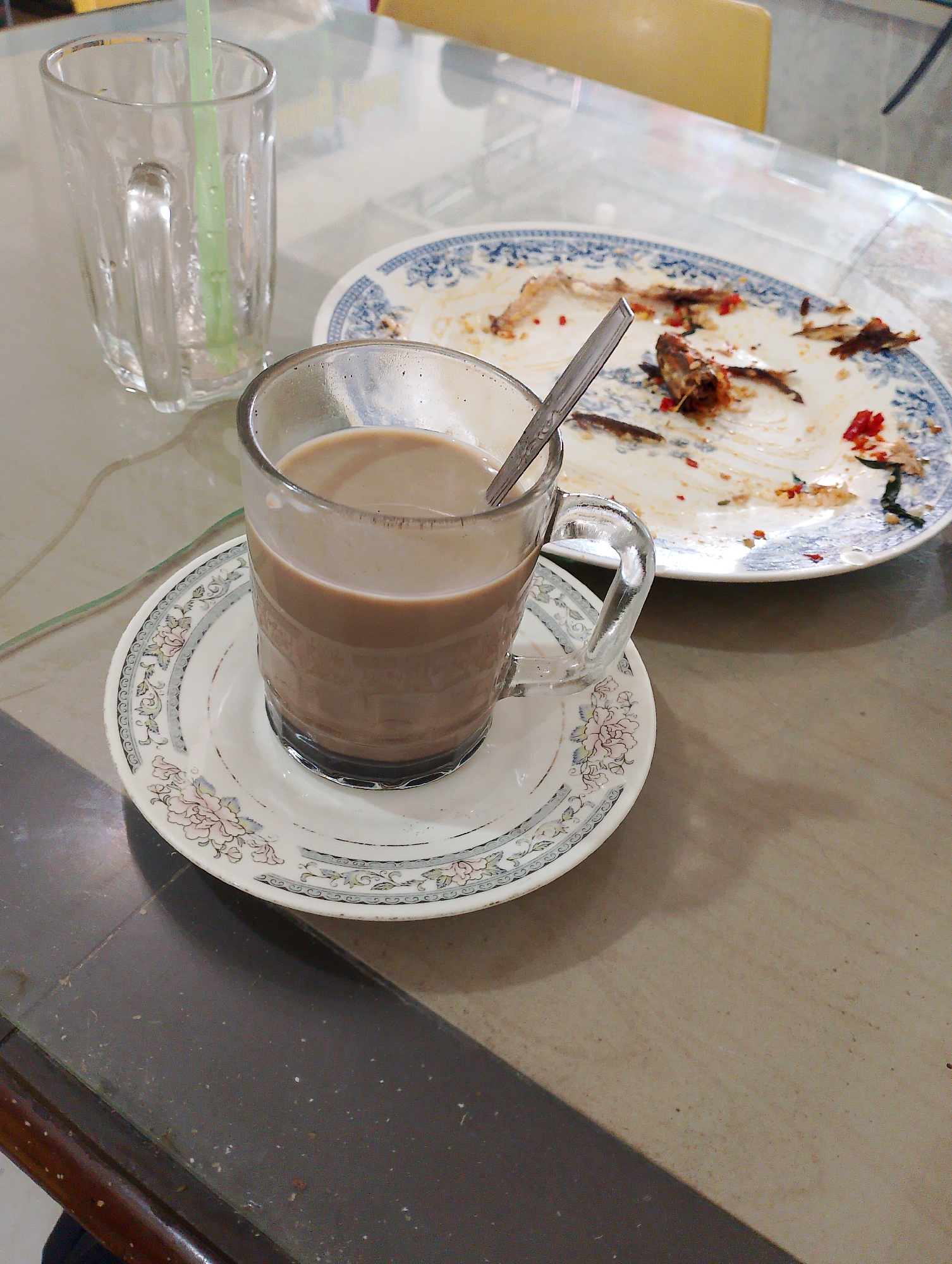 A close-up, high-angle shot shows a cup of coffee on a saucer, a used plate with fish bones, and an empty glass with a straw on a table. The coffee is in a clear glass mug with a handle, filled with a light brown liquid, and sits on a white saucer with a floral pattern. To the right, a white plate with a blue and white patterned rim is littered with the remains of fish, including bones and some red chili pieces. An empty, clear glass with a green straw stands to the left of the coffee. The table surface appears to be a smooth, mottled gray material, possibly a laminate or stone. The background shows the edge of a yellow chair and a reflective surface, suggesting an indoor setting, likely a restaurant or cafe. The scene is consistent with a meal that has recently been finished. The location is identified as Batam, Indonesia, suggesting this is a local eatery.