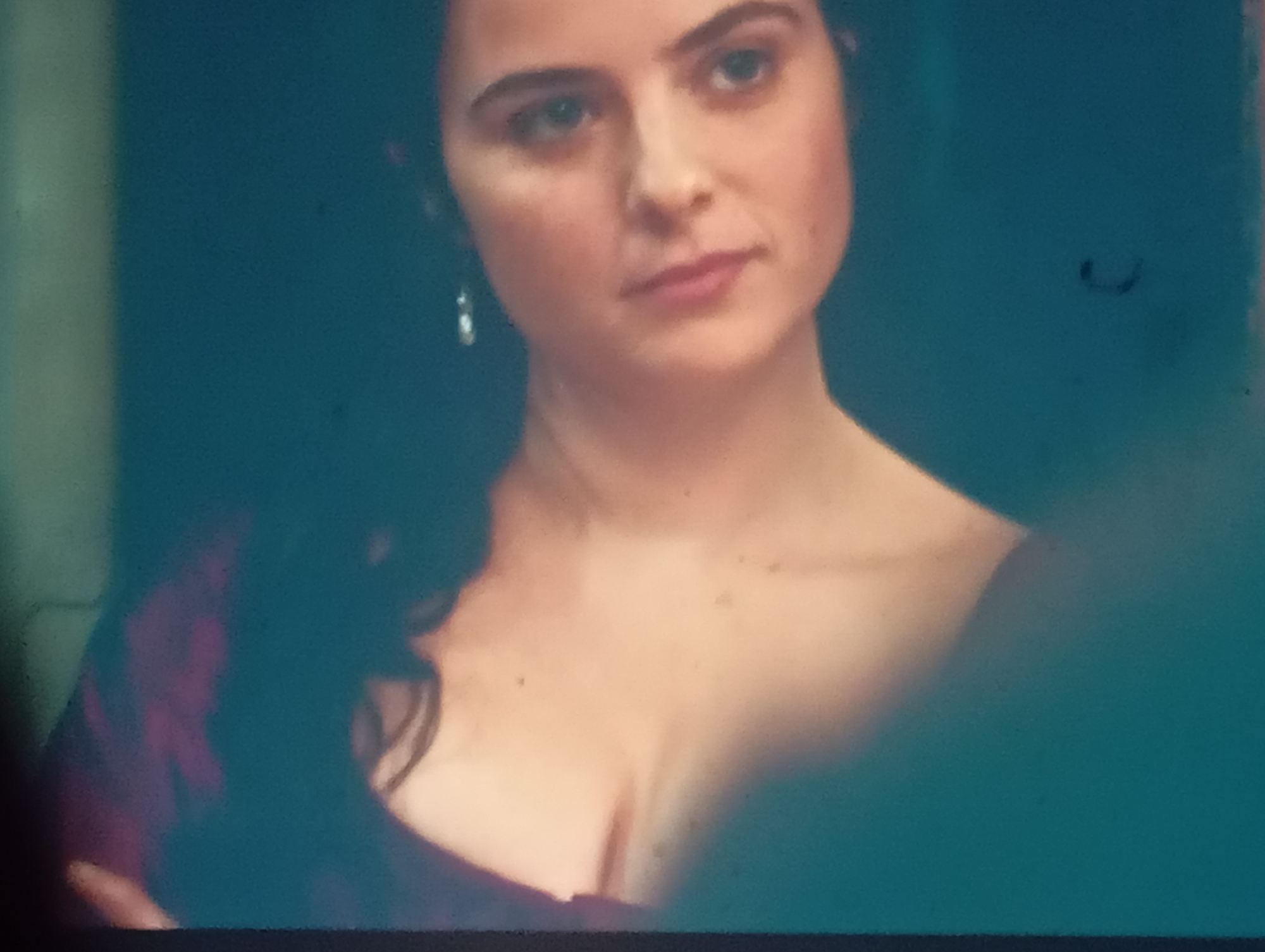 The image is a close-up, slightly out-of-focus shot of a young woman with dark hair and fair skin. She is wearing a dark purple dress with a visible neckline. Her left shoulder is bare, and she has a delicate earring in her left ear. The background is a dark, teal color, with some slight blurring. No specific details about Sofiivska Borshchahivka, Ukraine, can be discerned from this image.
