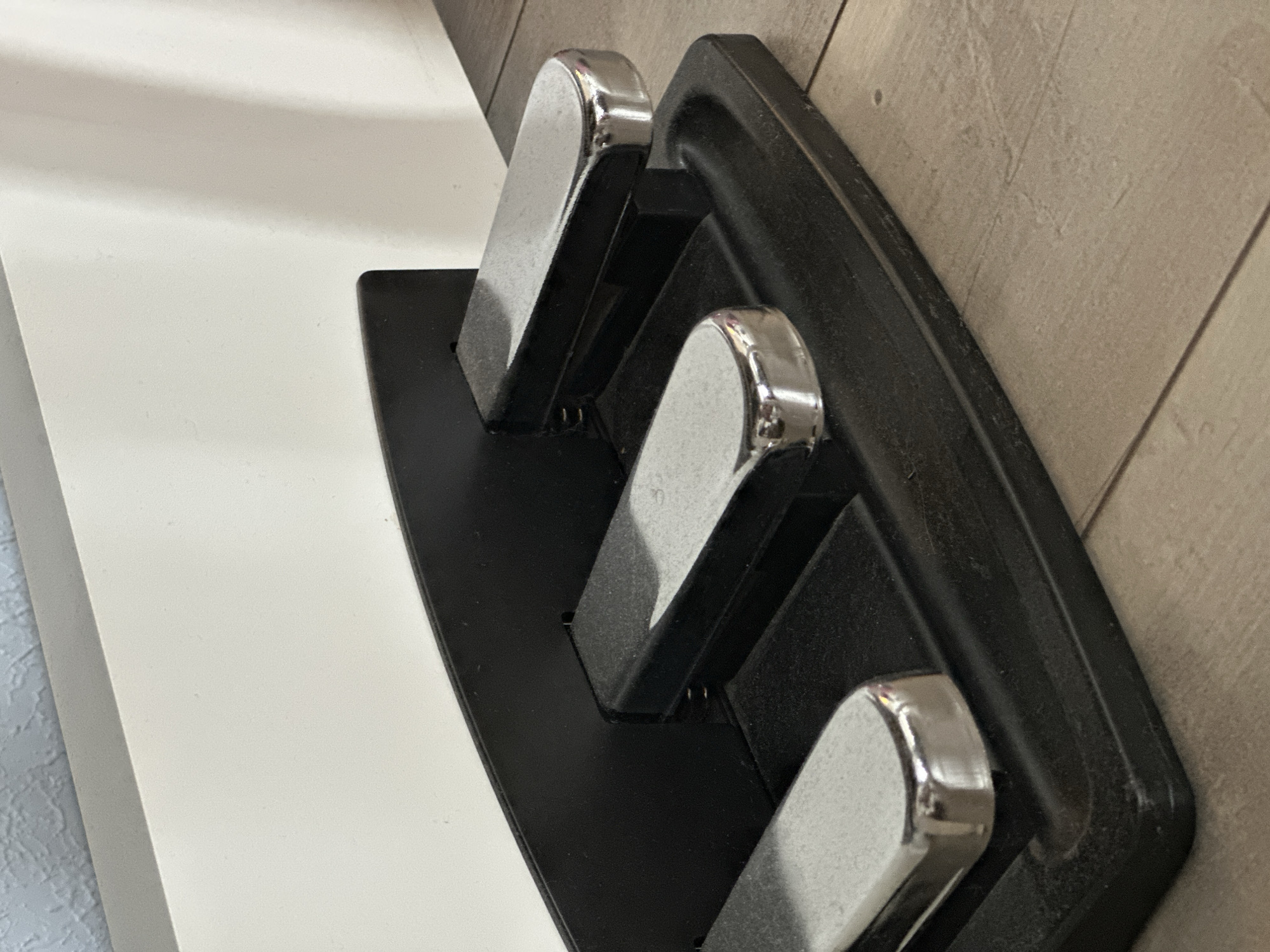 The image shows a close-up view of three piano pedals on a black stand, positioned on a wooden floor. The pedals are made of metal and have a reflective surface. They are arranged in a row, with the leftmost pedal partially visible. The stand has a curved black top and a flat base. In the background, a white surface is visible, possibly part of a piano or a wall. The lighting suggests an indoor setting, with shadows cast on the floor and the white surface. The scene is static and devoid of any human presence. The image does not contain any elements that would allow for its location to be identified as Nevinnomyssk, Russia.