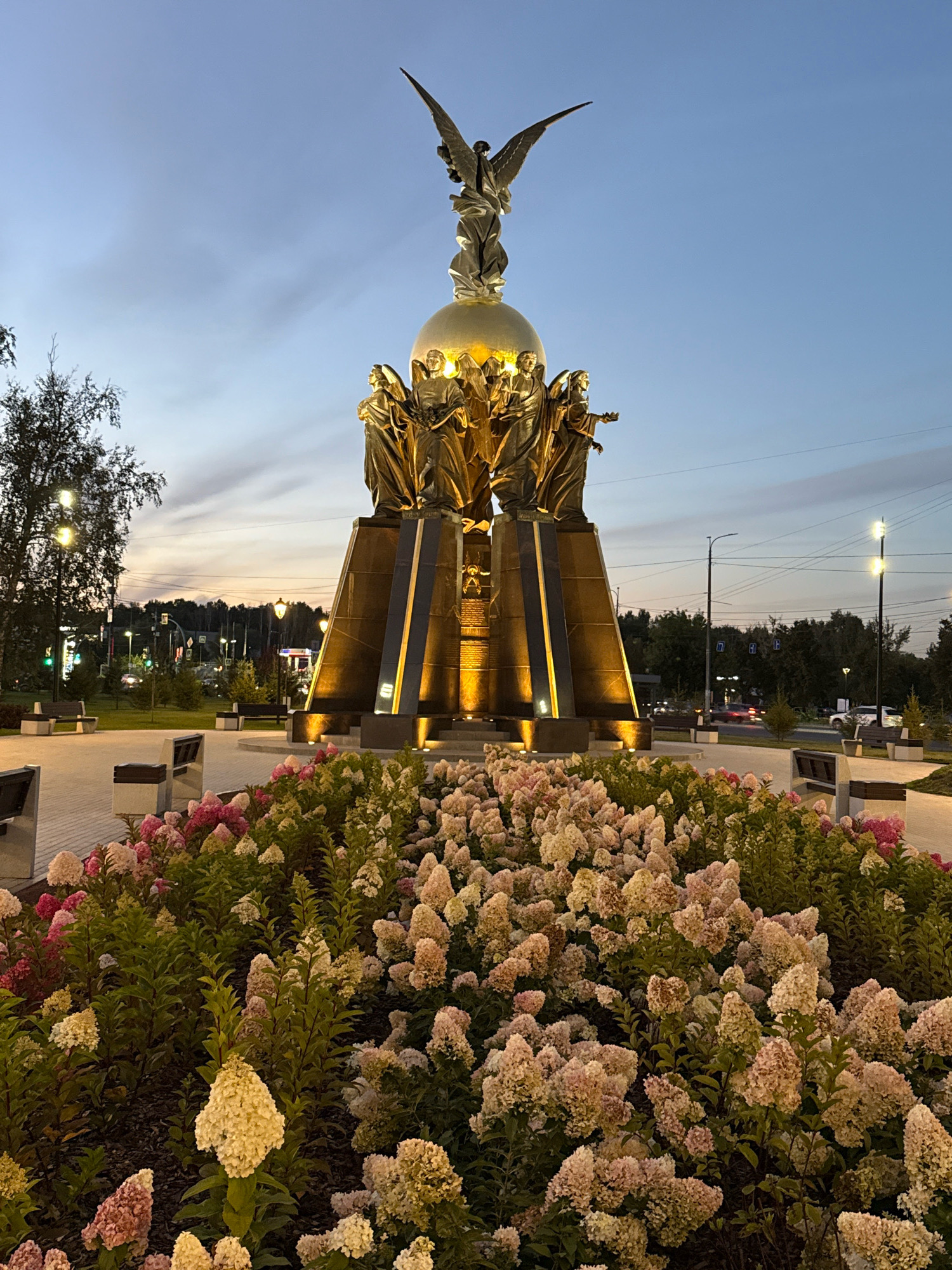The image depicts a monument at dusk in Chelyabinsk, Russia, located at the intersection of Komsomolsky Avenue and the road to the Chelyabinsk Tractor Plant. The monument features a golden globe at its apex, topped by a winged figure. Surrounding the globe are several golden statues of people. The base of the monument is a dark, stepped structure illuminated by spotlights. In the foreground, a flower bed is filled with hydrangeas in various shades of pink, white, and yellow. The background shows a tree-lined street with lampposts, benches, and passing cars. The sky is a twilight blue with some clouds.