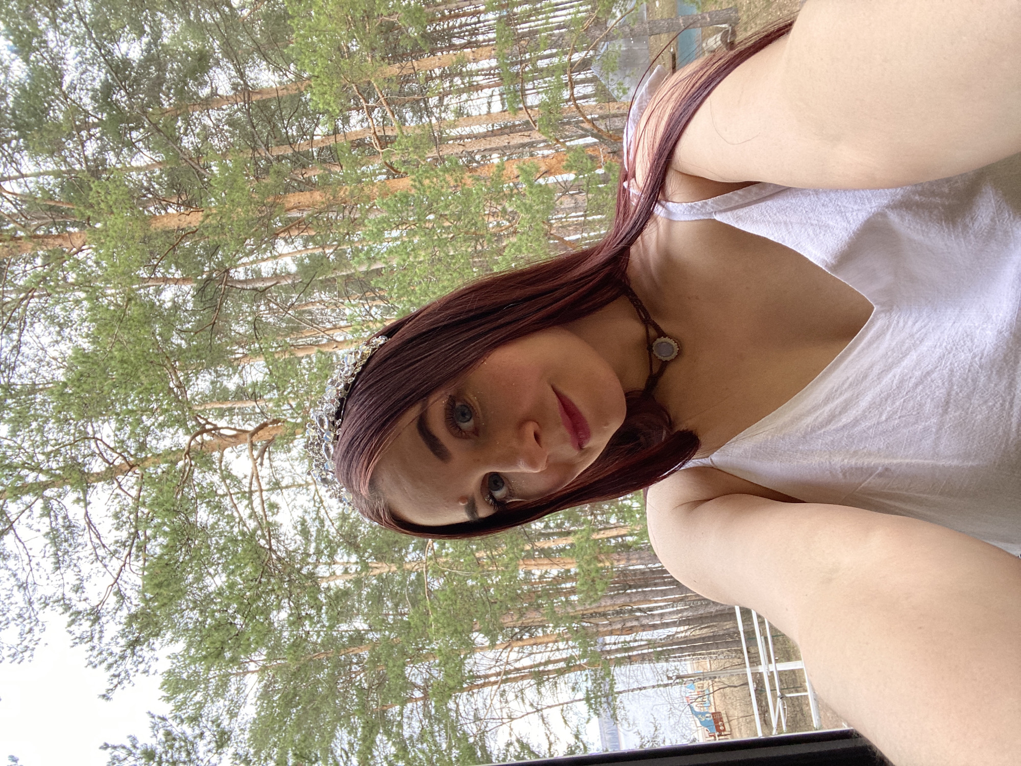 A woman with auburn hair and a jeweled headpiece takes a selfie in front of a wooded area. She is wearing a white top and a necklace with a stone pendant. Behind her are numerous tall trees with green foliage. The location is Chaikovsky, Russia.